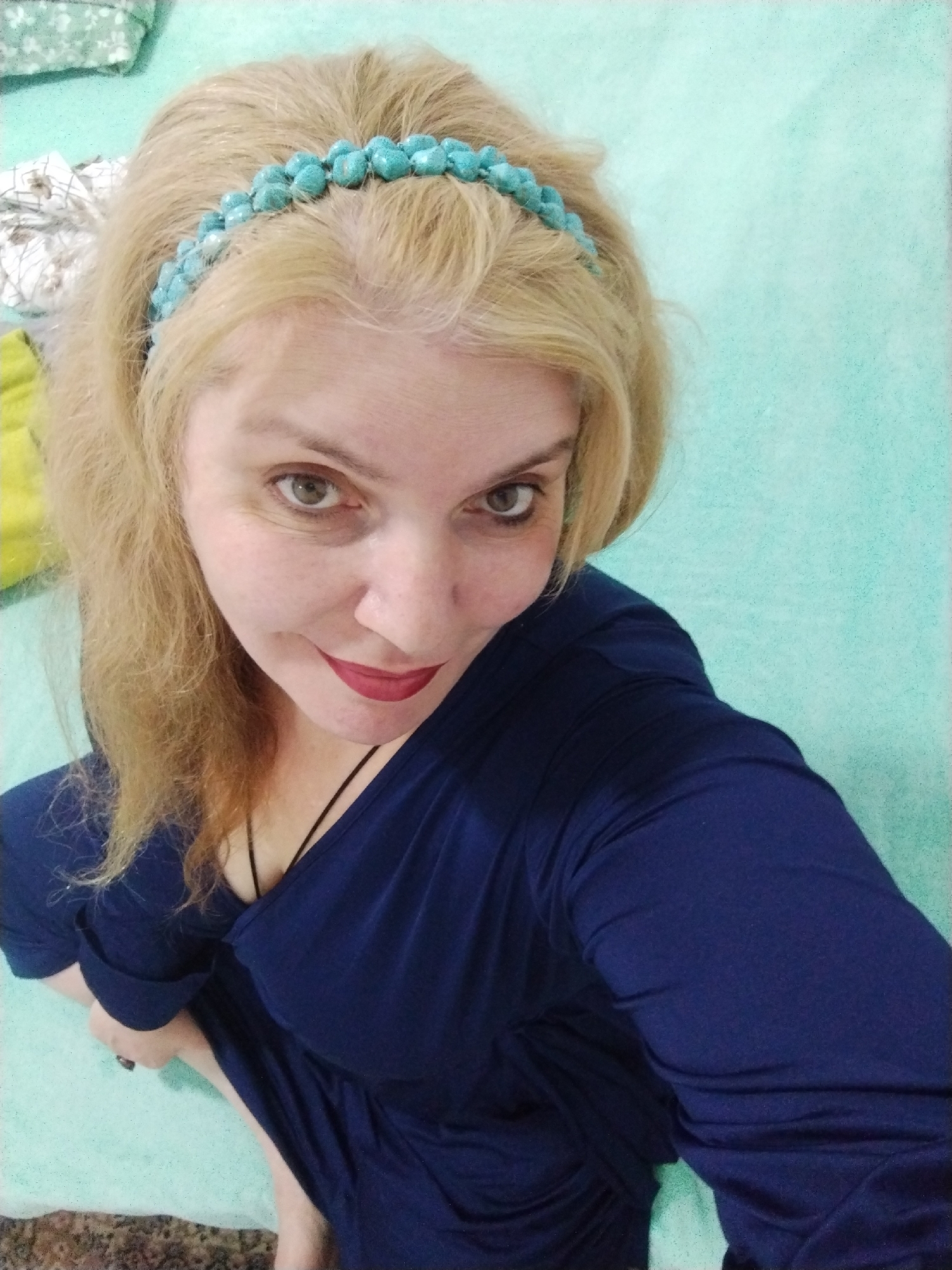 A blonde woman wearing a blue headband and blue dress sits on a light blue surface. She has fair skin, green eyes, and red lipstick. The background appears to be a room with a bed. The location, Al Mas'oudi, United Arab Emirates, is not visually evident in the image.
