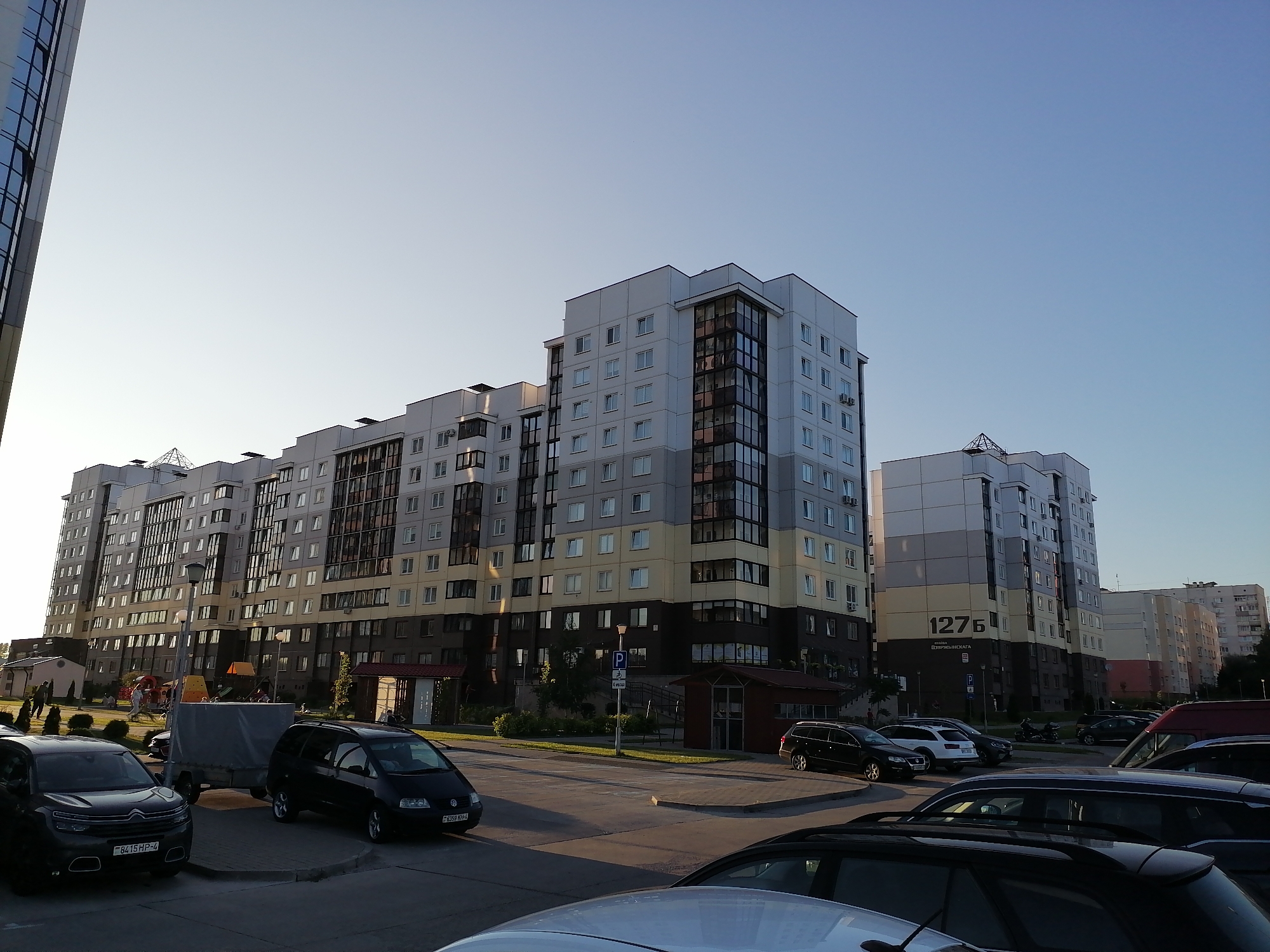 In Hrodna, Belarus, a wide shot captures a street scene dominated by a row of multi-story apartment buildings under a clear, light blue sky. The buildings are modern, with varying facade colors including white, grey, and yellow, and feature numerous windows and balconies. The apartment building in the center-right of the frame is prominently marked with the number "1276". A paved road and sidewalk are visible in the foreground, with several cars parked, including a dark grey Citroen, a black Volkswagen minivan, and a black SUV partially obscured. A grey tarp-covered trailer is parked behind the Citroen. To the left, a portion of a glass-fronted building is visible, reflecting the sky. The background reveals more apartment blocks stretching into the distance. A playground with colorful structures is visible near the base of the central apartment buildings. The lighting suggests it is late afternoon or early evening.