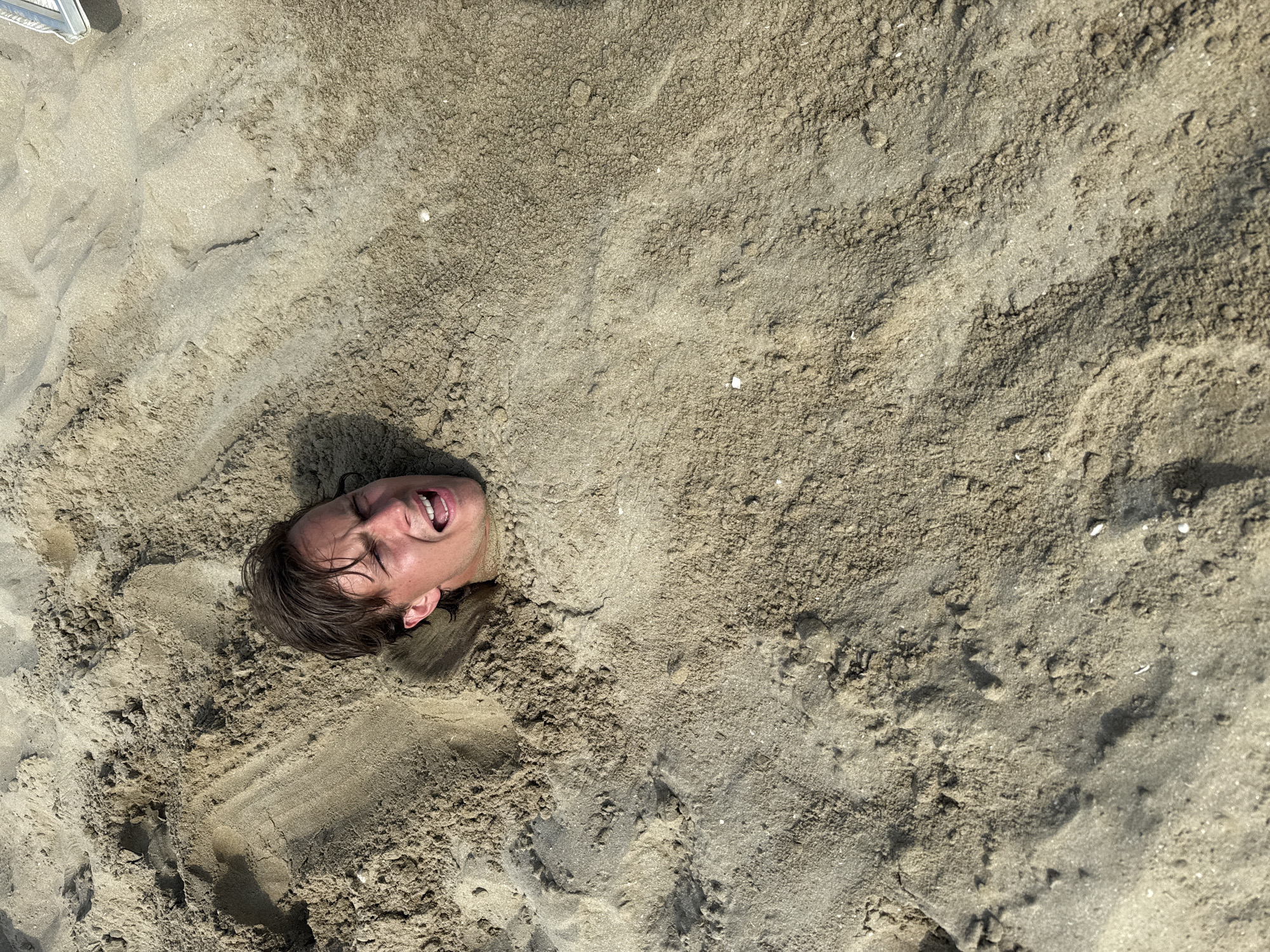 A young man with brown hair is buried in sand up to his neck, with his head and shoulders visible above the surface. His eyes are wide, and his mouth is open in a shout or gasp. The sand is light brown and textured, with visible grains and small shells. The scene appears to be a beach. The image is taken from a high-angle shot, looking down at the man's head. The background is mostly sand, with some shadows indicating the presence of other objects or people off-camera. This situation may be a playful prank on a beach, possibly in Rimini, Italy, known for its sandy shores and tourist activities.