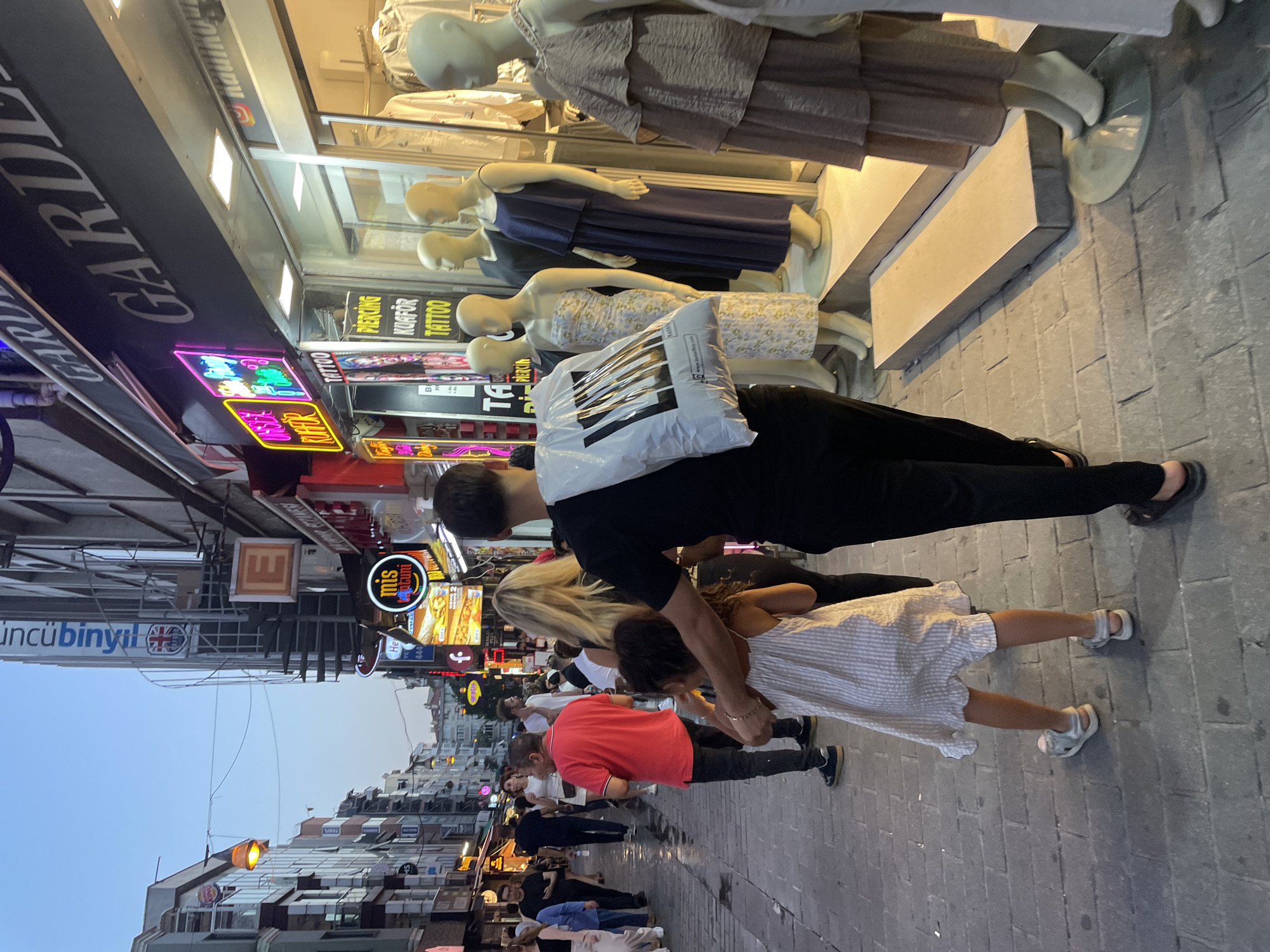 This media file depicts a street scene in Kadıköy, Turkey. The perspective is low, looking up at a street lined with shops and neon signs. Several people are visible, including a man in a black shirt and pants, a young girl in a striped dress and sandals, and other individuals walking and standing in the background. The man appears to be holding the girl's arm as they walk together.
The shops display mannequins dressed in clothing. Neon signs are prominent, with text in Turkish and English, including "GARD," "TATTOO," "PIERCING," "KUAFÖR," and "incübinyıl." The street is paved with cobblestones. The lighting suggests it is either late afternoon or early evening, with a warm, slightly hazy sky visible in the distance. The overall atmosphere appears to be one of bustling street life.