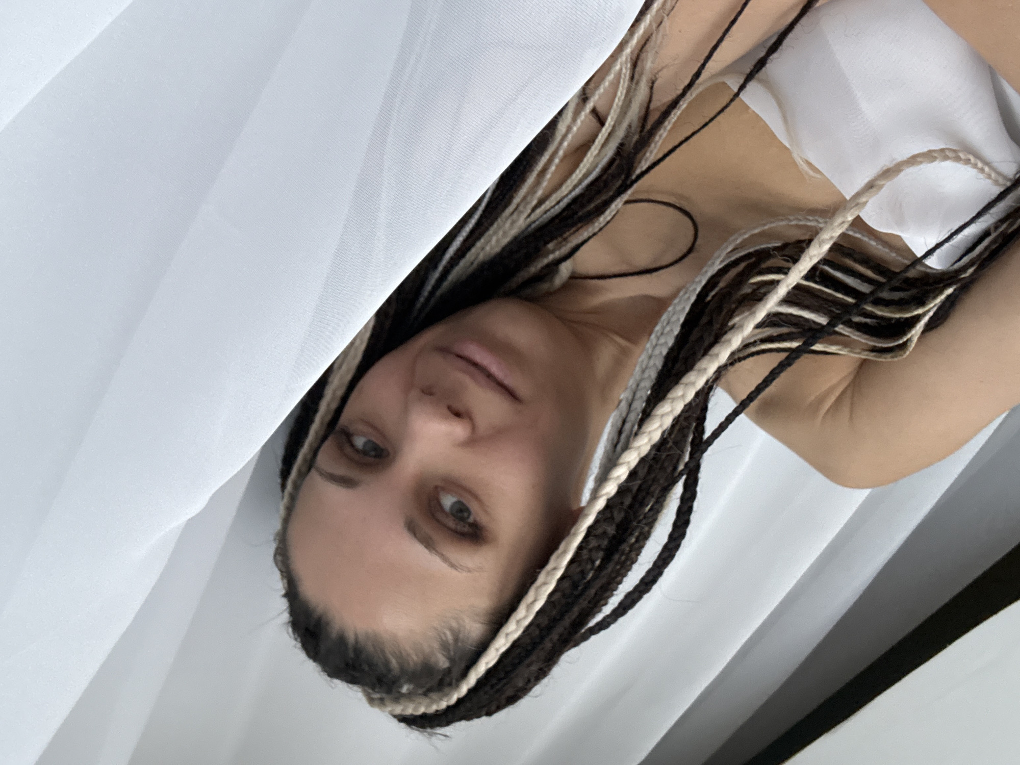 The media file displays a close-up portrait of a woman, oriented with her head to the left and body extending to the right, suggesting a 90-degree rotation from its original capture. She looks directly at the camera with a neutral, calm expression. Her hair is styled in numerous long, thin braids, featuring a prominent mix of dark brown, black, and light (blonde or white) strands. Her fair skin and a hint of a light-colored top are visible beneath what appears to be white fabric or drapery, which largely fills the background and foreground, creating soft folds. The lighting is even and diffused, characteristic of an indoor setting, likely during the day. The scene shows no specific activity beyond the woman posing. There is no discernible text within the image. The context for the image is Rusanovo, Russia.