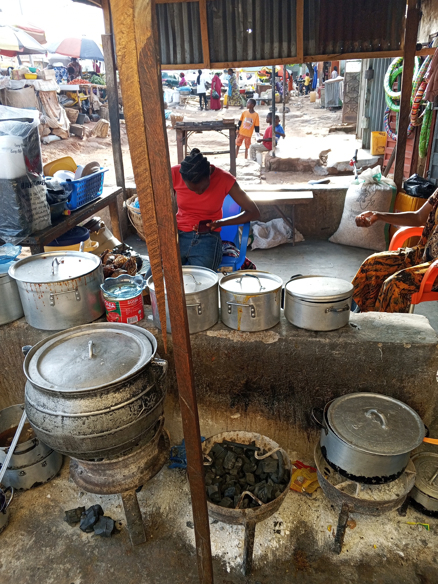 Here is a detailed description of the image:
**Location:** Gwong, Nigeria. The image appears to be inside a market stall or cooking area within a larger market.
**People:**
* Two women are visible. One is wearing a red shirt and jeans and is seated on a blue chair. The other is partially visible, wearing an orange patterned dress, and is seated in an orange chair.
* Several children are visible in the background.
* Other people are seen indistinctly in the crowded marketplace beyond the stall.
**Objects:**
* Multiple large metal pots of varying sizes are visible, some with lids on and some open. These appear to be cooking pots.
* A grill of fried meat.
* A can of "SONIA" tomato paste.
* Several charcoal grills with charcoal.
* Sacks of supplies or ingredients.
* A plastic blue basket.
* A bag of "Easy Life" plates.
**Actions:**
* One woman is sitting and seems to be using a phone.
* The second woman is sitting and holding a bean, perhaps sorting food items.
* Cooking appears to be taking place, indicated by the pots and charcoal fires.
**Interactions:** The main interaction is between the two women, presumably working together in the market stall. The wider interaction is with the customers in the market.
**Environment:**
* The scene is set in a covered stall or cooking area, possibly an extension of a more permanent structure.
* The area appears to be busy and somewhat cluttered, with various supplies and cooking equipment visible.
* The background shows a bustling marketplace with other stalls, people, and goods.
* The cooking area looks simple, possibly improvised, and could be a small business providing cooked food.