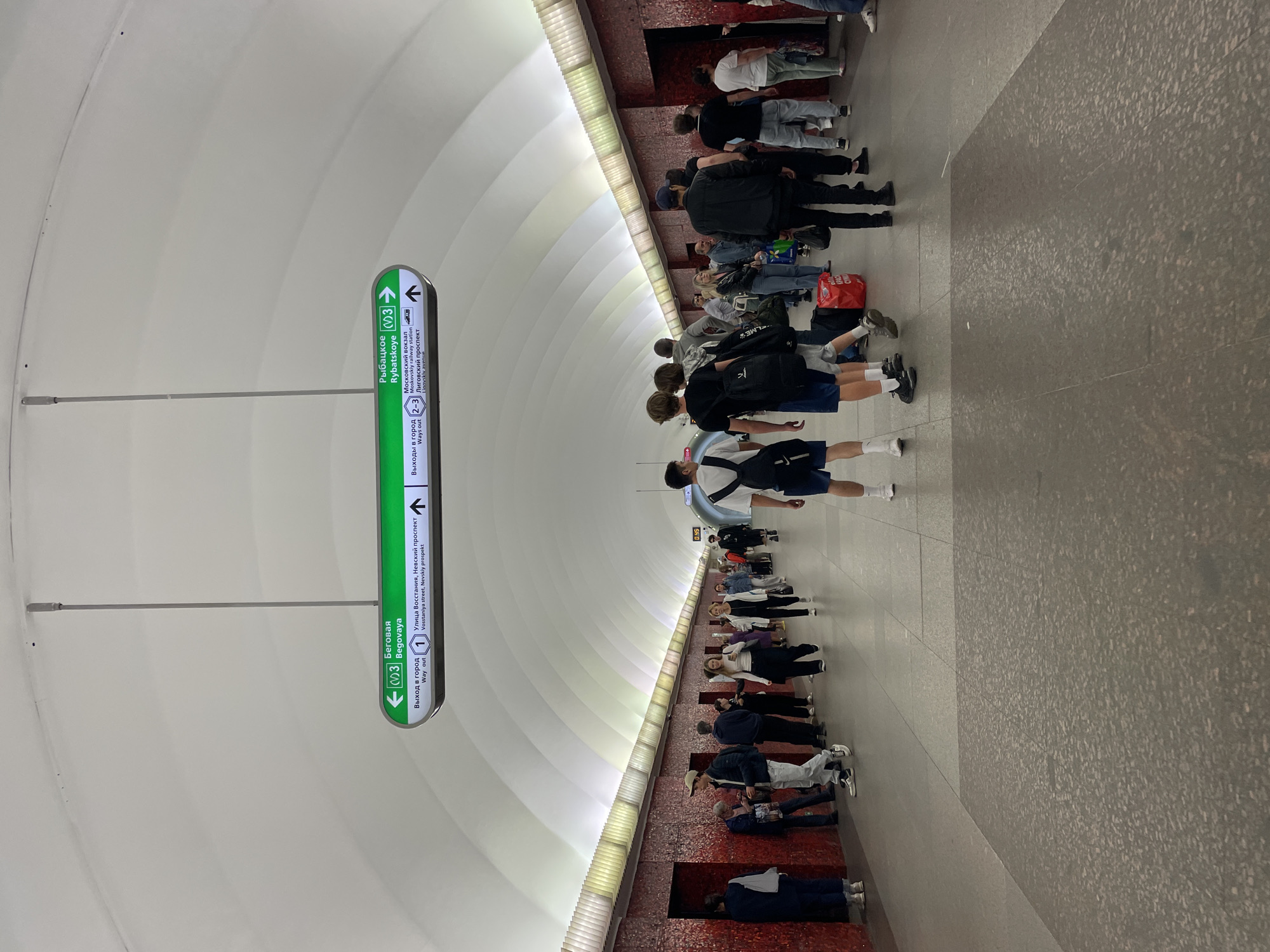 The image shows a view of a subway platform in an underground station. The platform is made of grey tiles, and the walls are covered in a red mosaic pattern. Above the platform, a white, arched ceiling spans the width of the station, illuminated by strip lighting. Several directional signs hang from the ceiling, with green backgrounds and white text indicating directions to different metro lines and exits, including "Begovaya," "Rybatskoye," "Voskstaniva street, Nevsky prospekt," and "Moskovskiy vokzal." A number of people are present on the platform, walking in various directions or standing. They are dressed in casual clothing. Some individuals are carrying bags or backpacks. The overall scene depicts a busy subway station environment.