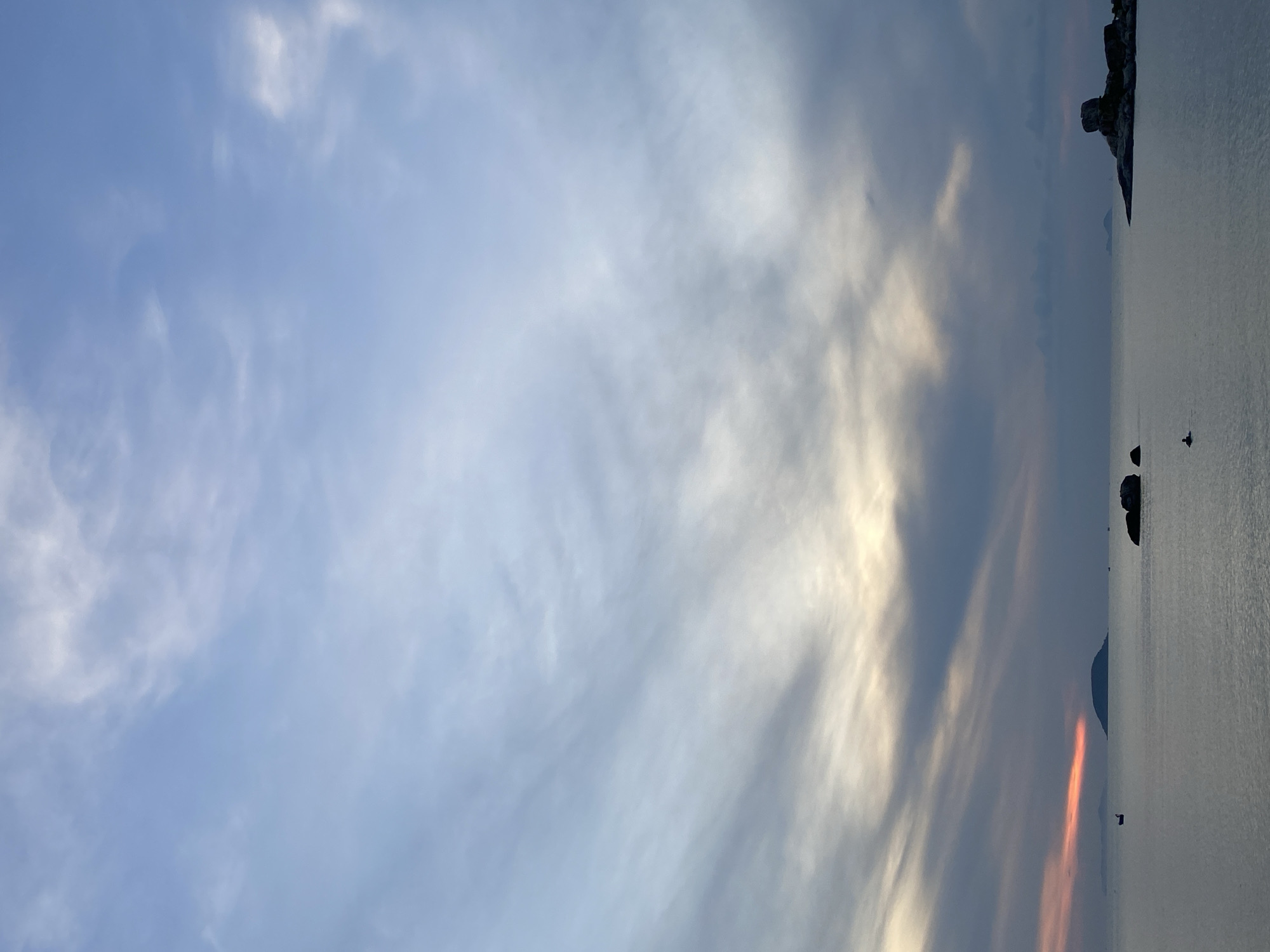 The image depicts a serene seascape at dusk, likely in Nha Trang, Vietnam, given the characteristic limestone karsts and calm waters. The sky is a gradient of soft blues and grays, with wisps of clouds catching the last rays of the setting sun, creating a gentle orange glow on the horizon. In the foreground, the calm sea stretches towards the horizon, reflecting the muted colors of the sky. Several dark, rounded rocks are scattered in the water, with one larger formation of rocks situated on the right side of the frame, appearing to jut out from a small island or landmass. In the distance, a small, dark island with a conical shape is visible to the left of center, and other smaller islands are faintly discernible on the horizon. A single, small boat with a dark silhouette is present in the water to the left of the conical island. On the right side of the image, a solitary figure can be seen in the water near a cluster of rocks, seemingly engaged in swimming or wading. The overall atmosphere is tranquil and contemplative, capturing a moment of natural beauty as day transitions into night.
