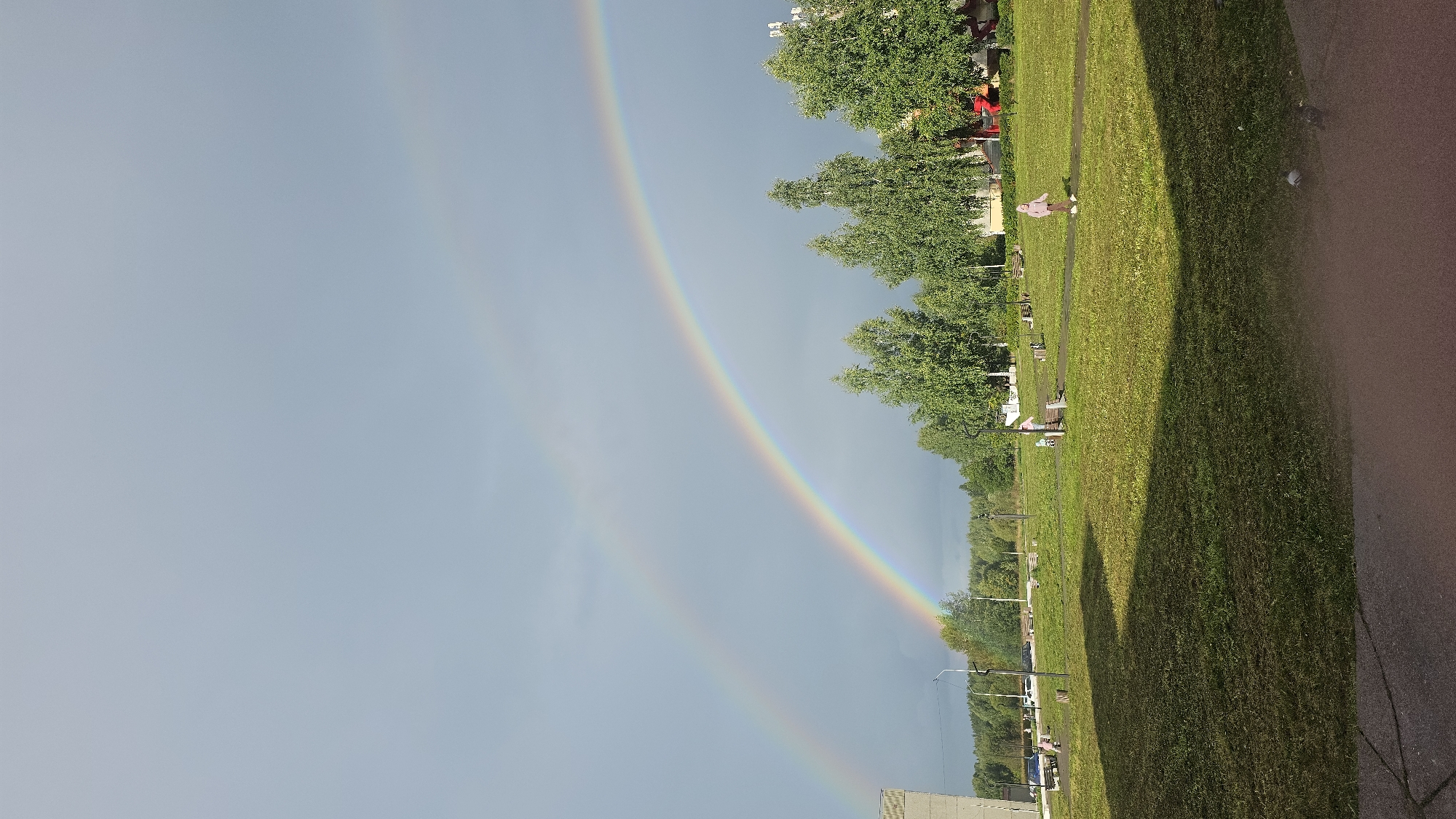 The image depicts a scene in a residential area of Markovskiy, Russia, following a rain shower. A double rainbow arcs across the overcast sky, with the primary rainbow being more vibrant than the secondary one. The foreground consists of a mown grass lawn, partially in shadow from an unseen structure. A paved walkway runs through the lawn. In the middle ground, several trees, some with sparse foliage and others with fuller canopies, are present. A few park benches are scattered throughout the area. On the walkway, a child wearing a pink hooded jacket and brown pants is running, and another child in a similar pink outfit is standing near a bench. A car is parked on the side of the road in the background, along with some buildings. The overall environment suggests a park or common area within a neighborhood.
