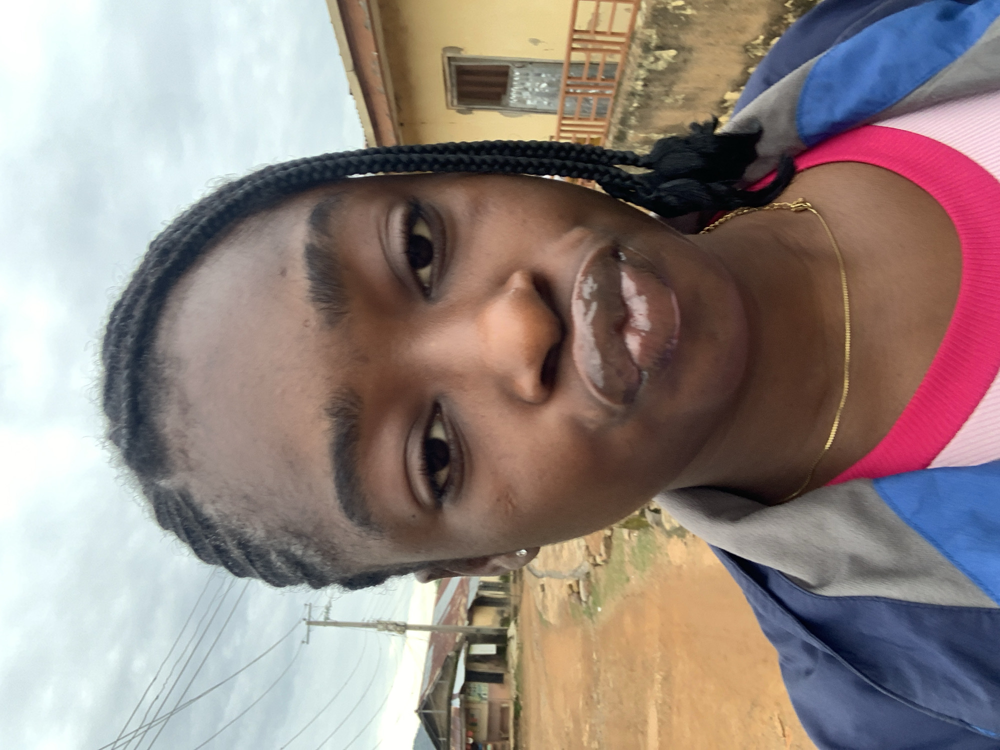 The image is a close-up selfie of a young woman with braided hair, wearing a blue, grey, and pink jacket over a pink top, and a thin gold chain necklace. She has prominent lips with a glossy sheen. In the background, there is a dirt road, some low-lying buildings with corrugated roofs, utility poles with wires, and a cloudy sky. The setting appears to be a rural or semi-urban area, possibly a residential street, consistent with the appearance of Rafin Par, Nigeria. The overall lighting suggests an overcast day.