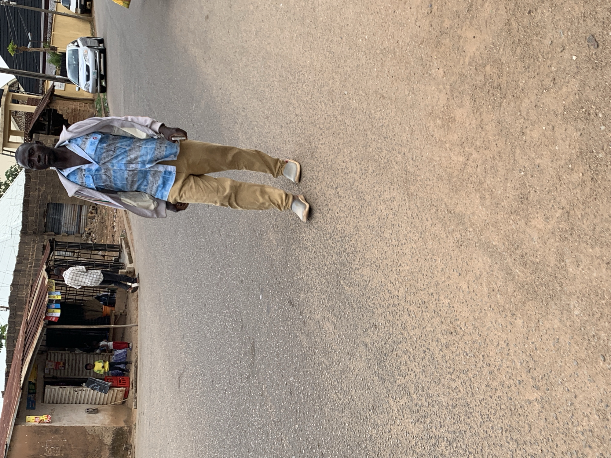 An African man wearing a blue patterned shirt and beige trousers walks down a paved street in Jos, Nigeria. He appears to be holding a light-colored cell phone in his right hand. The man is facing the camera. Behind him on the left is a small shop with various items on display, and a couple of other people are visible in the shop area. A white vehicle is parked further up the street. The street itself is slightly angled, receding into the distance on the right. The sky overhead is overcast.