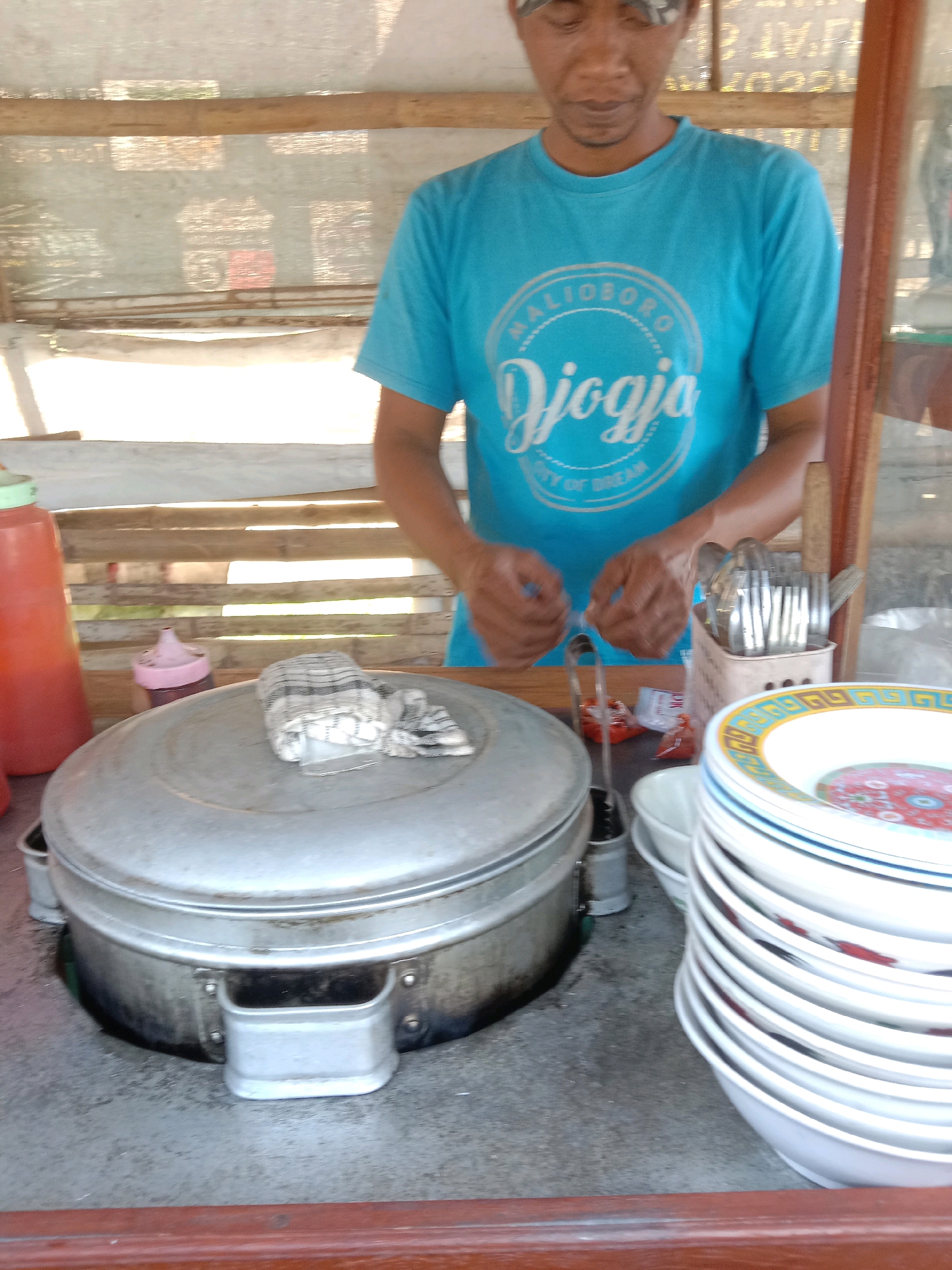 In Kedungrejo, Indonesia, a man in a blue t-shirt is standing behind a food stall. He is wearing a headscarf. On the stall, there are several items including a large metal pot with a lid covered by a cloth, a red bottle, a pink bottle, a basket of cutlery, and a stack of white bowls with colorful rims. The background shows a bamboo structure and a clear plastic sheet. The man appears to be preparing food.