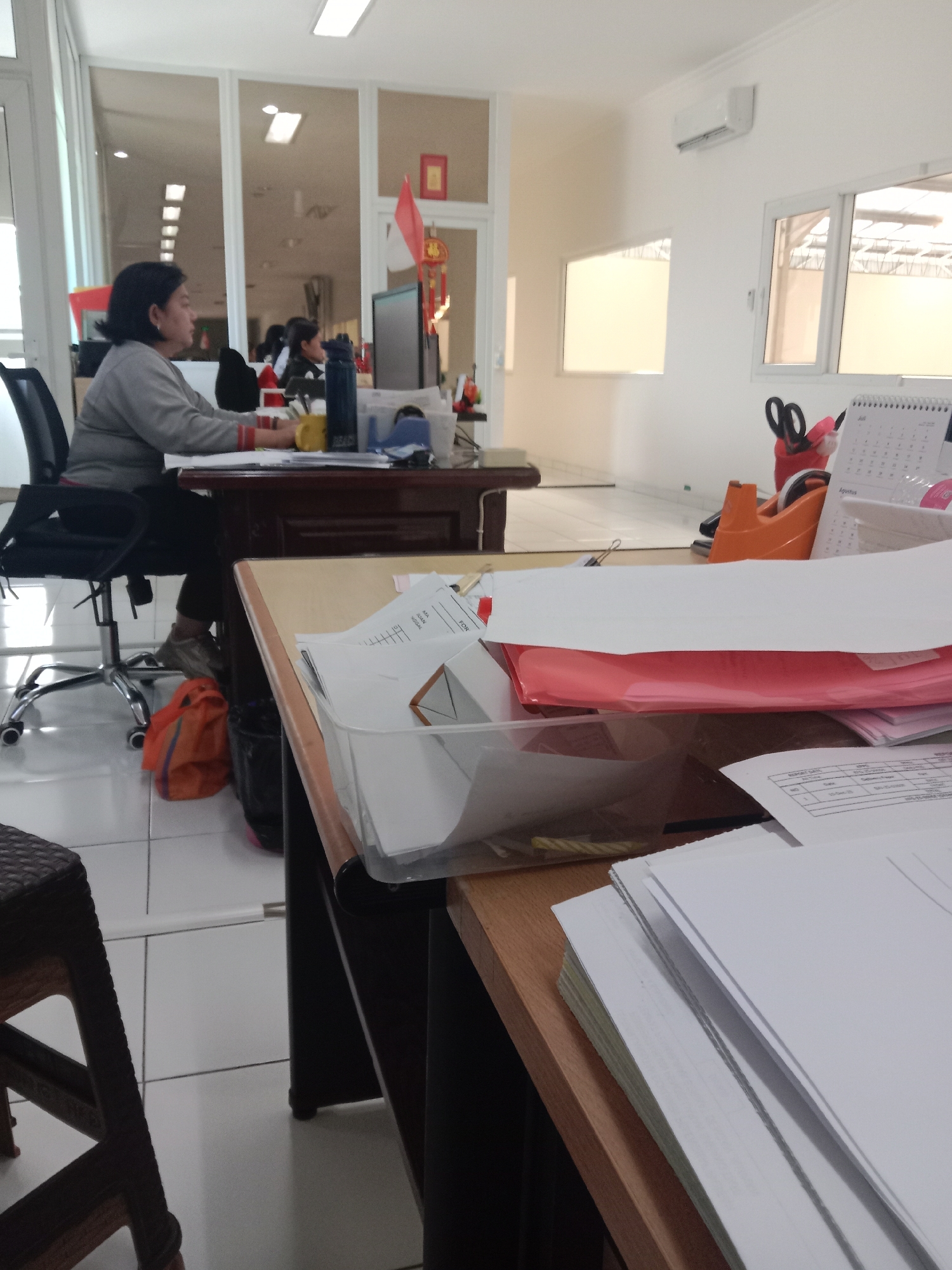 The image shows an office environment, likely in Semarang, Indonesia, with a woman working at a desk. The woman, with dark hair, is seated in an office chair and appears to be typing or interacting with a computer. She is wearing a grey long-sleeved shirt and dark pants. Her workspace includes a computer monitor, a yellow mug, a blue tape dispenser, and various papers and folders. An orange bag is visible on the floor near her chair. The office has tiled flooring, white walls, and large windows. In the foreground, another desk is laden with stacks of papers, a clear plastic tray containing more documents, a red folder, and a desk calendar that reads "Agustus" (August). The overall impression is one of a functional, somewhat cluttered office space.