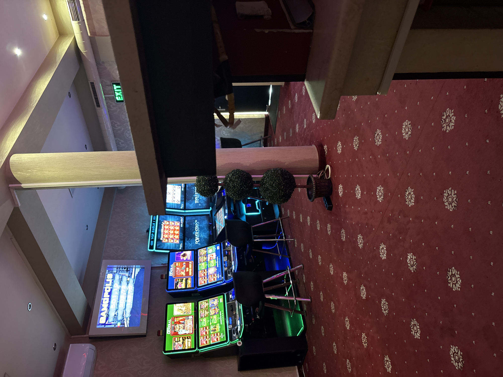 This image depicts the interior of a casino in Karlovo, Bulgaria. The room is carpeted with a red patterned rug. Several slot machines are visible, with screens displaying game graphics and numbers. Two of the machines are the "General" brand, with one displaying "VIP Eagle 1" and the other "Power Stars." Another machine is labeled "Triple Effect." A large screen mounted on the wall displays the word "GAMOPOLIS" and several numerical values. An illuminated "EXIT" sign is mounted on the ceiling. Two bar stools are placed in front of two of the slot machines. A decorative plant with three spherical sections is positioned in the foreground, partially obscuring a portion of the scene. The lighting appears to be a mix of ambient and artificial light, with some areas illuminated by the slot machines themselves. The environment suggests a typical casino setting, with rows of gaming machines and an emphasis on visual stimulation through light and graphics.