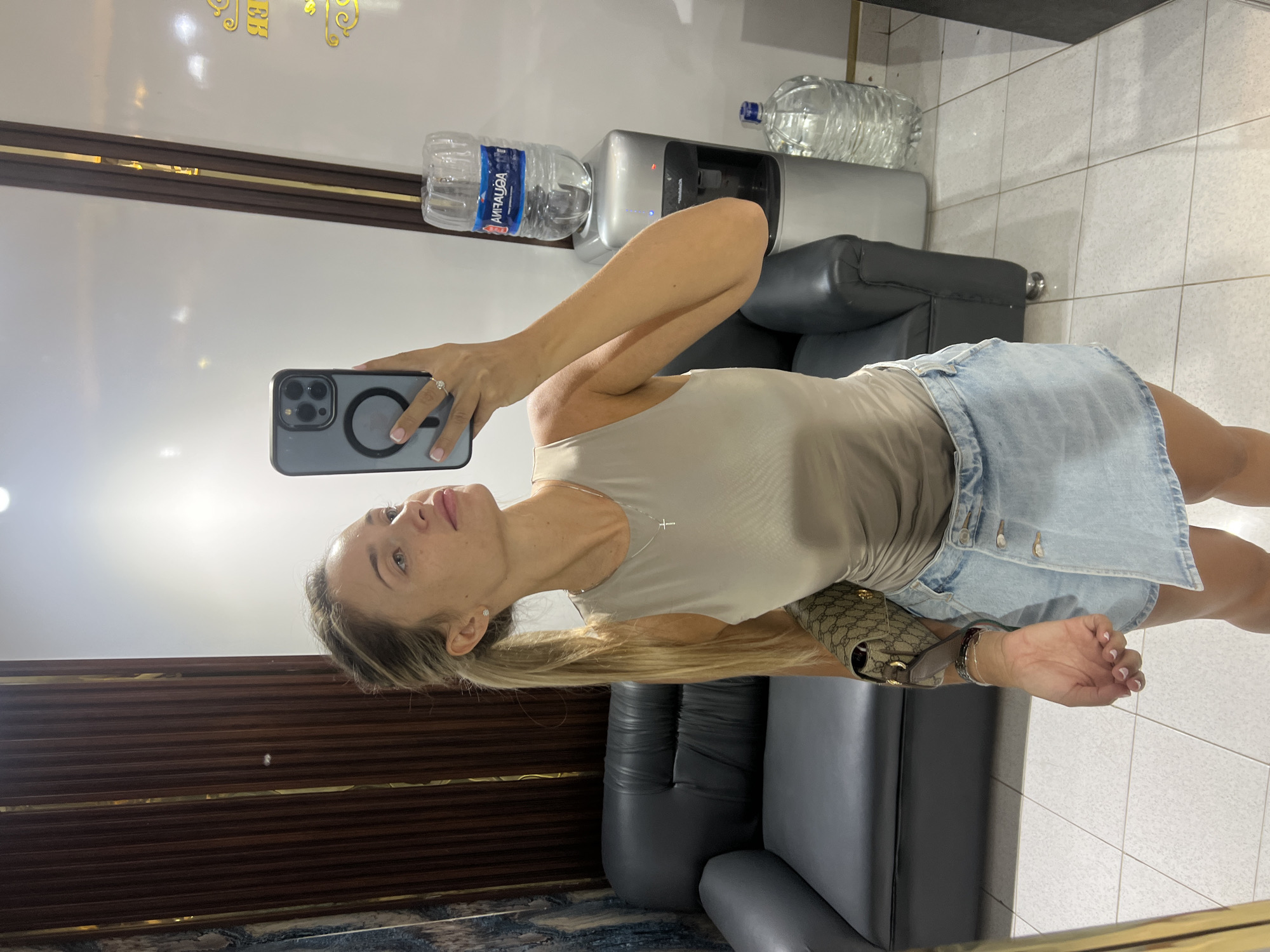 A woman is taking a mirror selfie in what appears to be a hotel room or a salon. She has blonde hair pulled back in a ponytail and is wearing a beige tank top and a denim skirt. She is holding a smartphone to take the picture and is wearing a ring on her finger. In the background, there is a black sofa, a water cooler with a bottle of water on top, and a large bottle of water on the floor. There is also a wooden partition and a mirror. The floor is tiled. The image was taken in Sharm El Sheikh, Egypt.