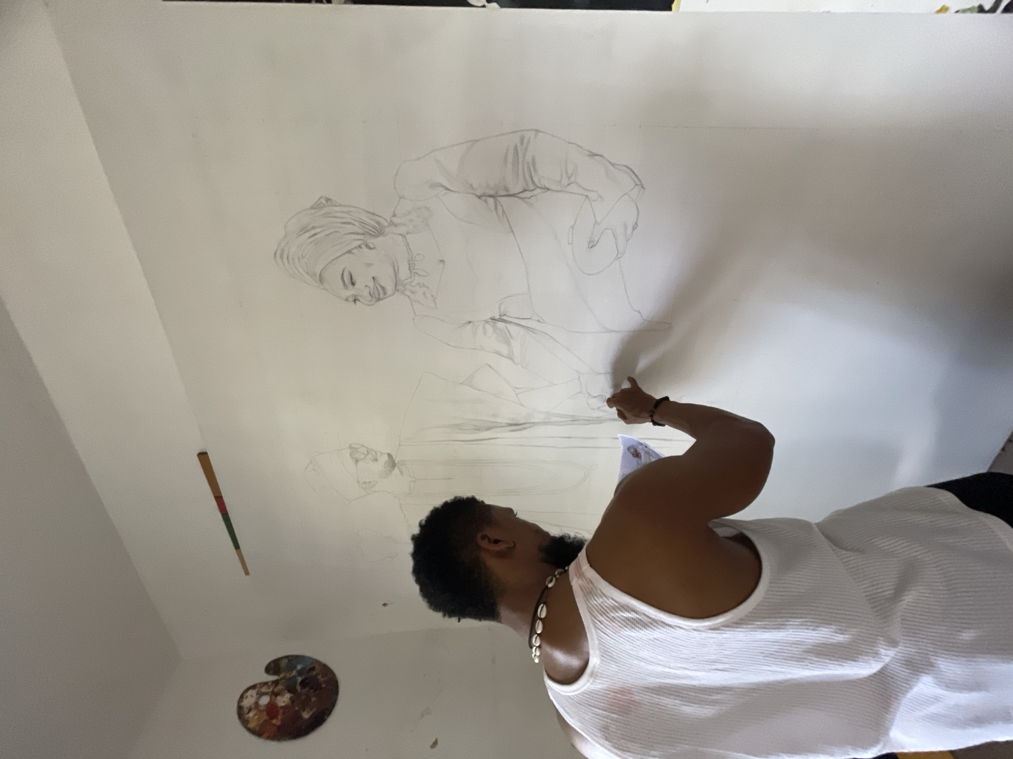 A man is drawing on a large sheet of white paper attached to a white wall. He is standing, wearing a white ribbed tank top and a black beaded necklace. The drawing on the paper depicts two figures in traditional clothing. A paint palette is affixed to the wall below the drawing. A paint brush is affixed to the wall near the top left corner of the drawing. The scene takes place inside a white-walled room. There is no other information to indicate that the location is in Ado, Nigeria.