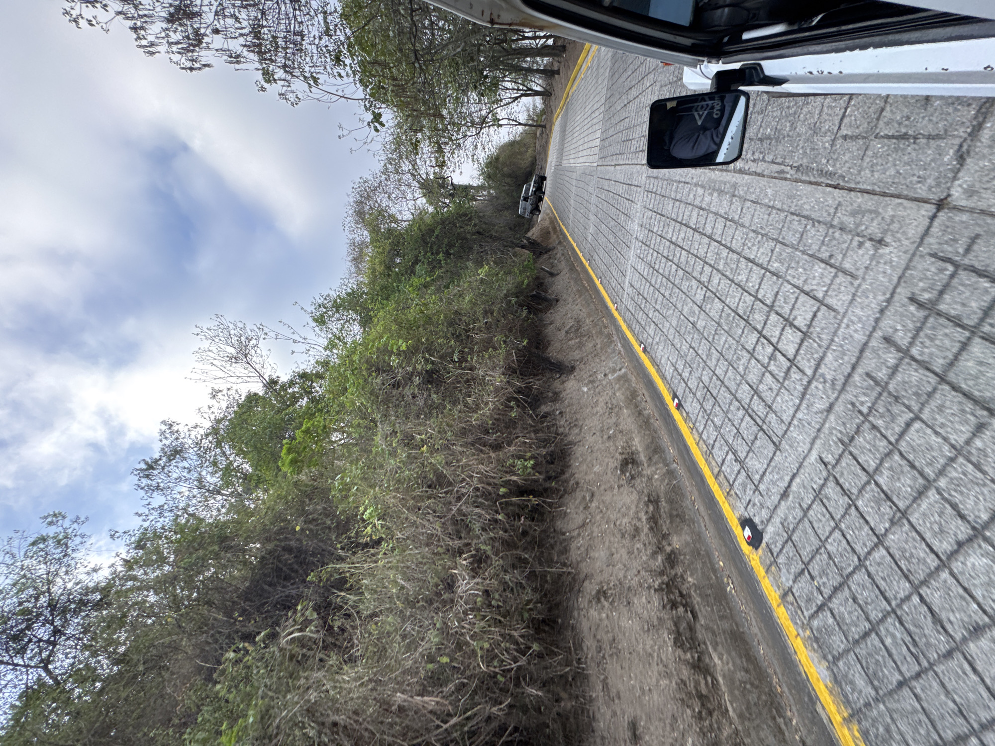 The image shows a road with cobblestone paving in Caracas, Venezuela. The road has a yellow line marking the edge and small reflective markers. A hillside covered in dense vegetation borders one side of the road. The sky is visible with scattered clouds. A rearview mirror is visible in the top right corner. Another vehicle is ahead on the road. The photo was likely taken from inside a moving vehicle.