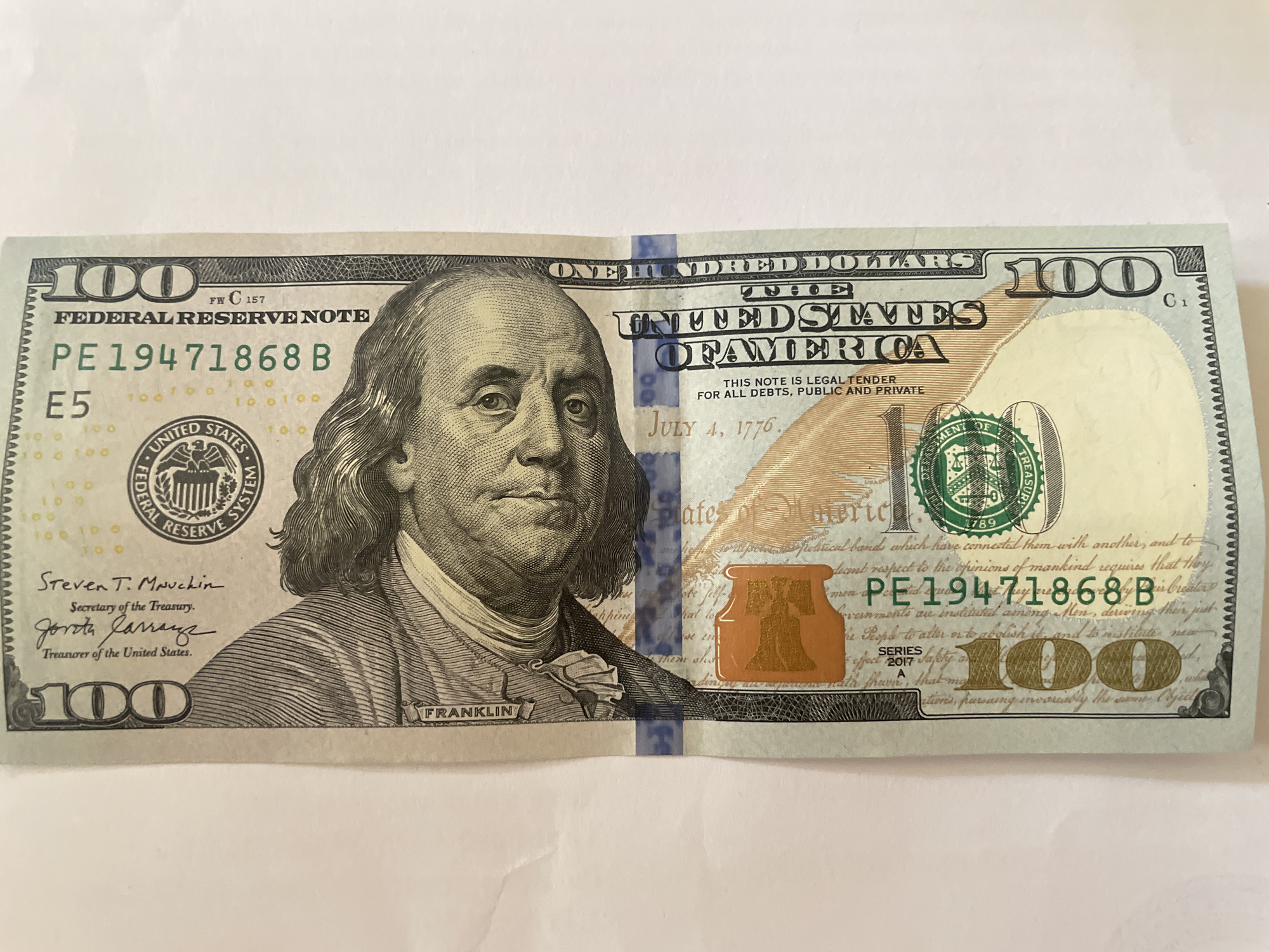 A United States $100 bill, featuring Benjamin Franklin, is photographed on a white background. The bill is oriented vertically and slightly creased in the center. The serial number PE19471868B is clearly visible. The seal of the Federal Reserve System and the Treasury Department are also visible. The image does not depict any location in the city of Minsk, Belarus, or any other geographical location.