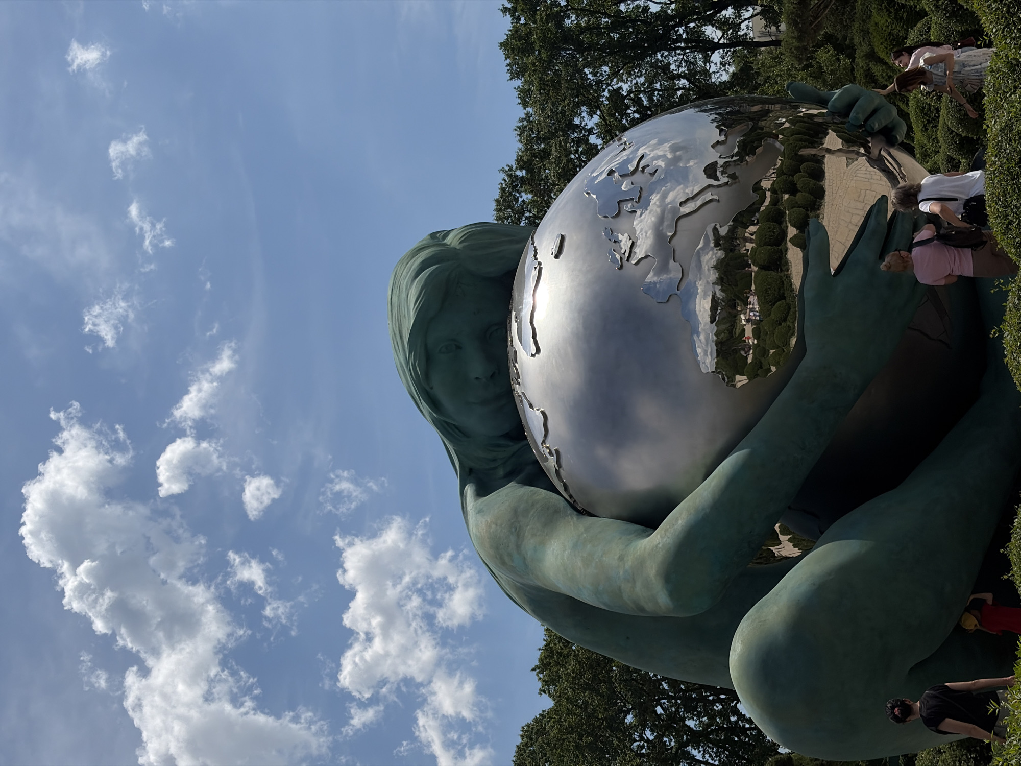The image displays a monumental sculpture titled "The Embrace" by Dashi Namdakov, situated within Galitsky Park (also known as Krasnodar Park) in Krasnodar, Russia, under a clear blue sky dotted with white cumulus clouds.
The primary subject is a colossal, patinated bronze sculpture of a female figure, exhibiting a prominent teal-green hue indicative of weathering. The figure is depicted in a kneeling or crouching posture, her head resting on a large, highly polished spherical globe crafted from reflective metal, likely stainless steel. Her arms encircle and cradle the globe. The globe's surface features a raised, metallic relief map of continents, with Europe, Africa, and parts of Asia clearly discernible. The globe acts as a mirror, reflecting the blue sky, white clouds, surrounding green trees, and various elements of the park's landscape, including paved pathways and distant park visitors.
The surrounding environment is characterized by vibrant greenery. Tall, mature deciduous trees with dense foliage are visible in the background on both the left and right sides of the frame. The immediate foreground and the base of the sculpture are densely covered with well-maintained green bushes and ground cover.
Several park visitors are present in the foreground, observing the sculpture. On the far left, an adult woman with dark hair tied back, wearing a black top and dark bottoms, stands facing towards the right. A child in a red t-shirt is partially visible beside her. On the right side, a group of four adult women is observing the artwork. Two women, seen from behind, stand closer to the globe: one wears a light pink t-shirt and a dark backpack, and the other wears a white t-shirt with a dark backpack. To their right, a woman in a light-colored, patterned dress stands, looking towards the sculpture. Another woman is partially visible on the far right edge of the frame.
The scene is brightly illuminated by daylight, with direct sunlight causing prominent reflections and highlights on the metallic surface of the globe.