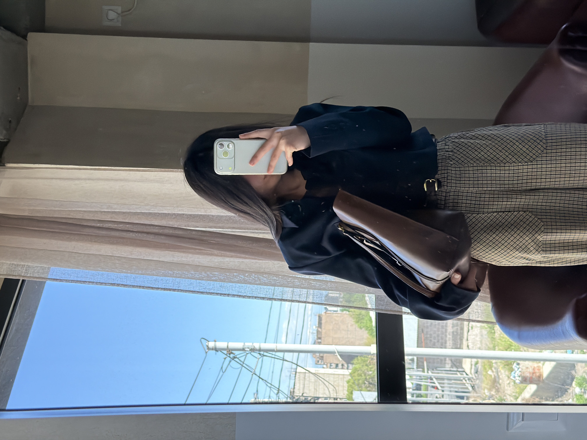 A person is taking a selfie in a mirror. The person is wearing a black jacket, a brown and beige plaid skirt or pants, and holding a brown purse. Outside the window, scaffolding, buildings, and trees are visible under a bright blue sky. Given the context of Yerevan, Armenia, the buildings are likely typical Soviet-era or post-Soviet construction. The presence of scaffolding suggests ongoing construction or renovation in the area.