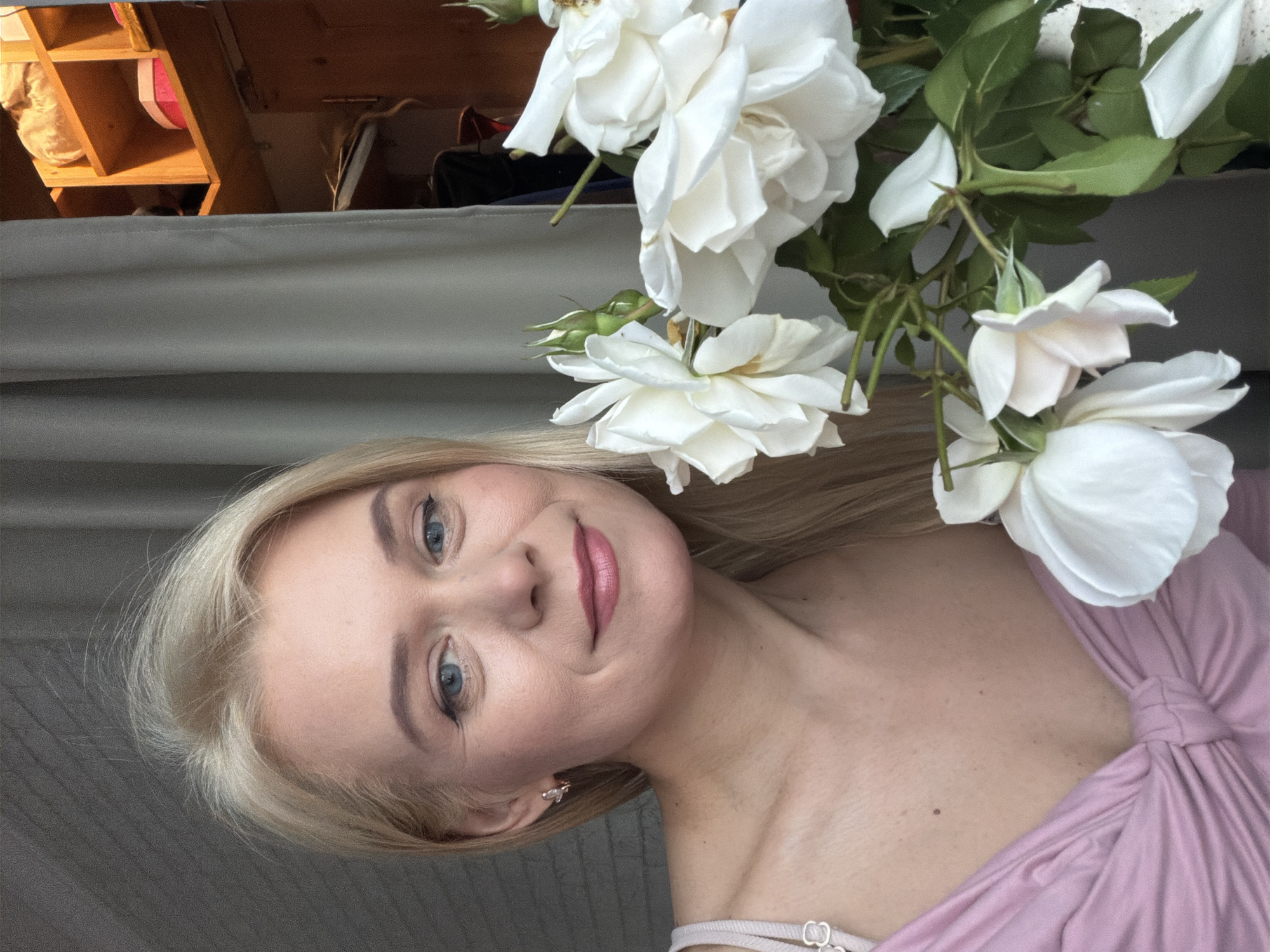 A young Caucasian woman with blonde hair and blue eyes is looking up at the camera, a slight smile on her face. She is wearing a pink dress with a knotted detail across the chest. A cluster of white roses with green leaves is in the upper right corner of the frame, appearing to be held or placed near her. The background is out of focus, but suggests an indoor setting with shelving units visible in the top left corner, and a curtain or fabric draped behind the woman. The lighting appears to be natural, possibly from a window, casting a soft glow on her face. The image conveys a sense of gentle beauty and perhaps a moment of peaceful contemplation. The time of day is not explicitly indicated, but the lighting suggests it could be daytime. No visible text is present in the image.