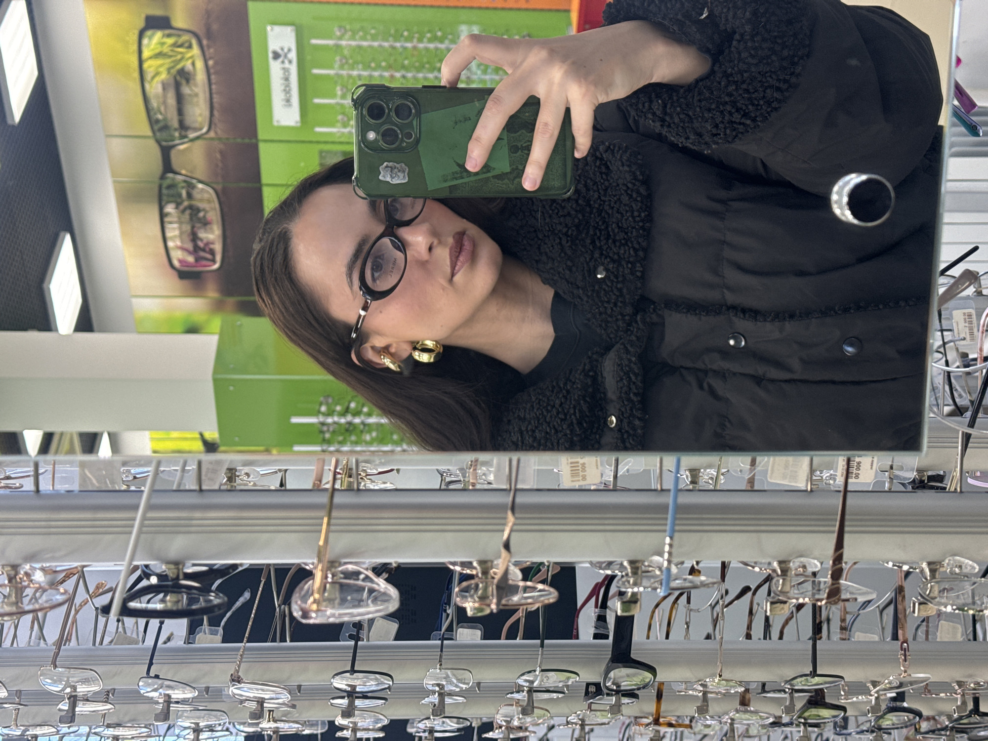 A woman is taking a selfie in a mirror. She has long dark hair, wears glasses and gold hoop earrings. She is wearing a black coat with a fluffy collar. She holds a green iPhone in front of her face. The reflection in the mirror also shows a display of eyeglasses. The location appears to be a store selling eyeglasses in Krasnoyarsk, Russia, based on the signage visible in the background.