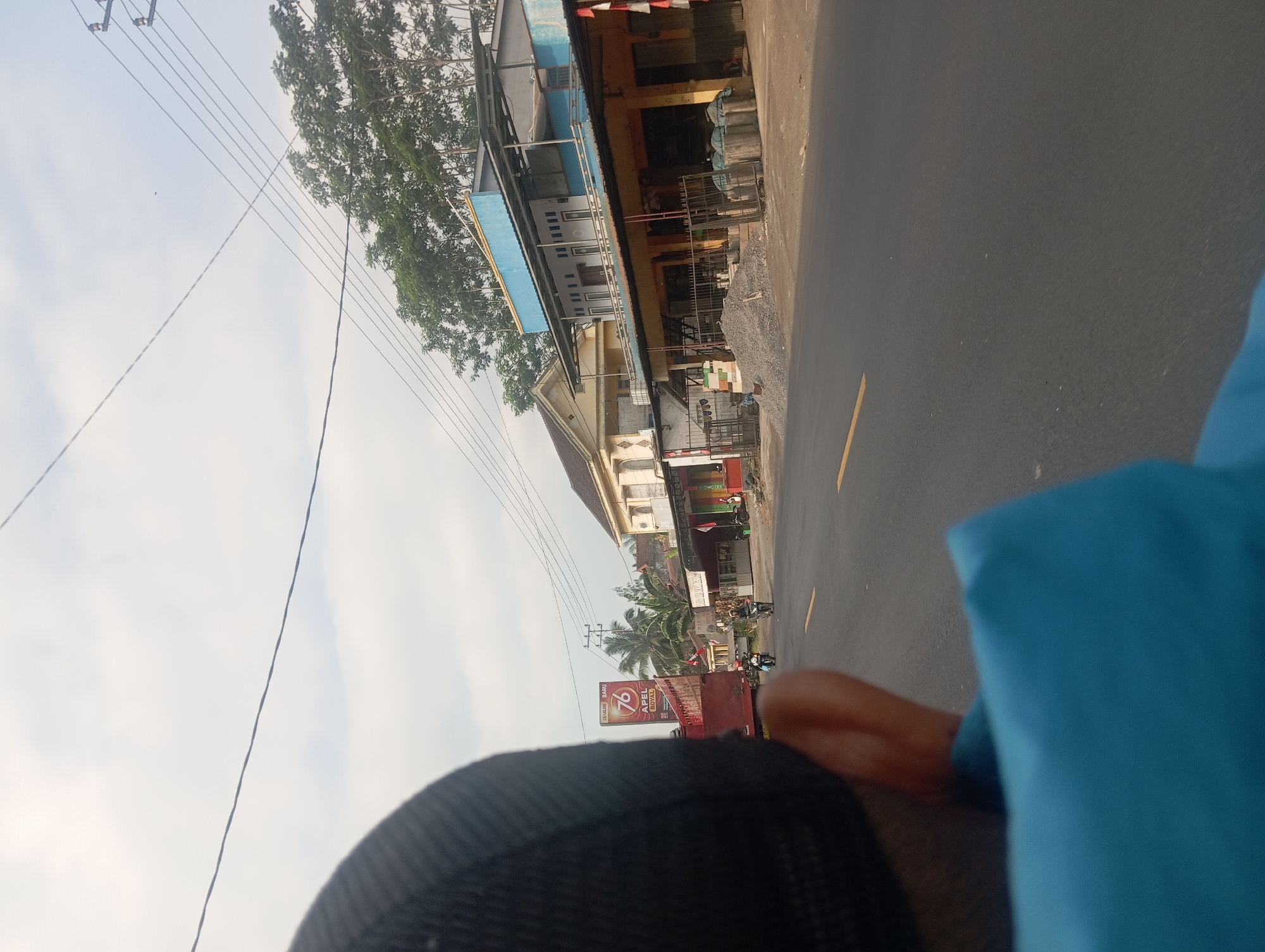 The image depicts a street scene in the city of Klakah, Indonesia, viewed from the perspective of someone riding in a vehicle. In the foreground, a person's head wearing a black cap and their blue clothing are visible, suggesting they are a passenger. The road is asphalt with a yellow line marking. On the right side of the road, there are several buildings with shops on the ground floor. One building has a blue and white upper story. In front of the shops, there are piles of gravel, metal cages, and cylindrical objects. Further down the street, there are two motorcycles with riders. To the left, a large red truck is partially visible, and behind it, a tall billboard for "Djarum 76 APEL ROYAL" stands. Palm trees and other lush green trees line the background, with a cloudy sky overhead. The scene appears to be a typical daytime street in a rural or semi-urban area.