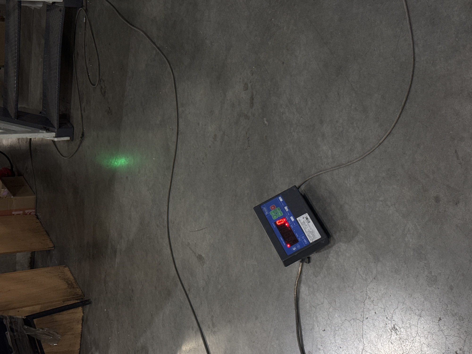 A gray concrete floor is the primary visual element. A black digital scale with a red LED display sits on the floor, connected to a power cord. A green laser dot is projected onto the floor to the left of the scale. Staircases and boxes are partially visible on the left side of the image. The specific location (Perm, Russia) is not discernable from the image. There are no people in the scene.