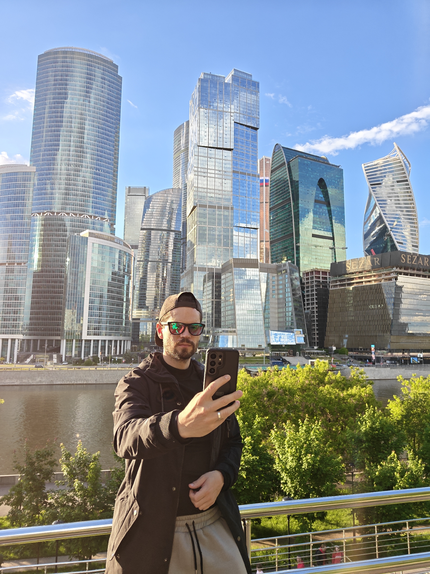 A medium shot captures a man in front of the Moscow International Business Center, often called Moscow City, in Moscow, Russia. The man, wearing a black jacket, gray sweatpants, a black t-shirt, a cap, and red-tinted sunglasses, is holding a smartphone. He appears to be taking a photo or video. The skyscrapers of Moscow City rise in the background, reflected in the river. A row of trees lines the riverbank in front of the buildings. A metal railing separates the man from the scenery. The sky is blue with scattered clouds.