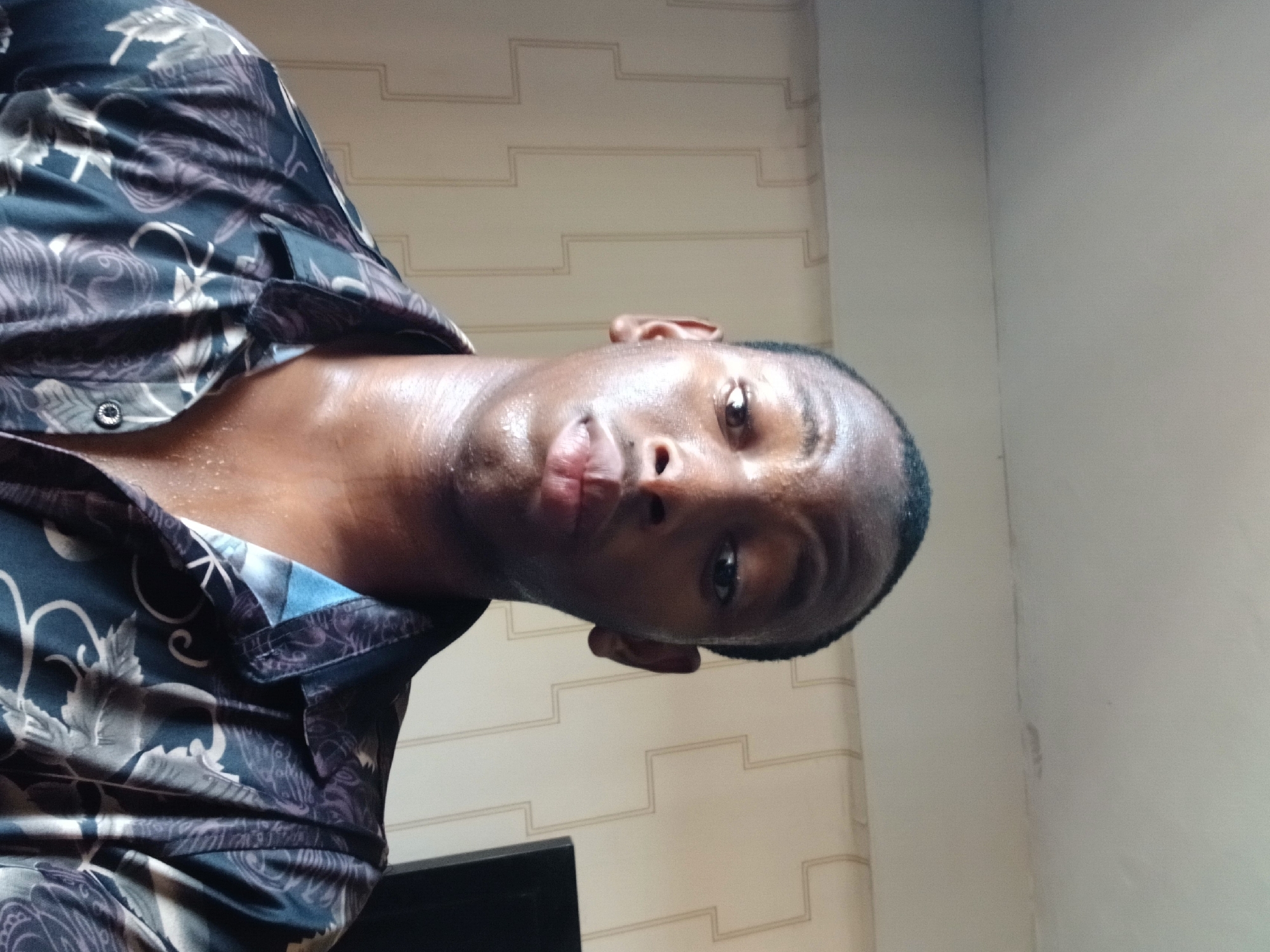 A man with dark skin and a shaved head is wearing a patterned shirt. The background consists of a patterned wall. The image was likely taken indoors. It is not possible to provide the location in Lagos, Nigeria from the given image.