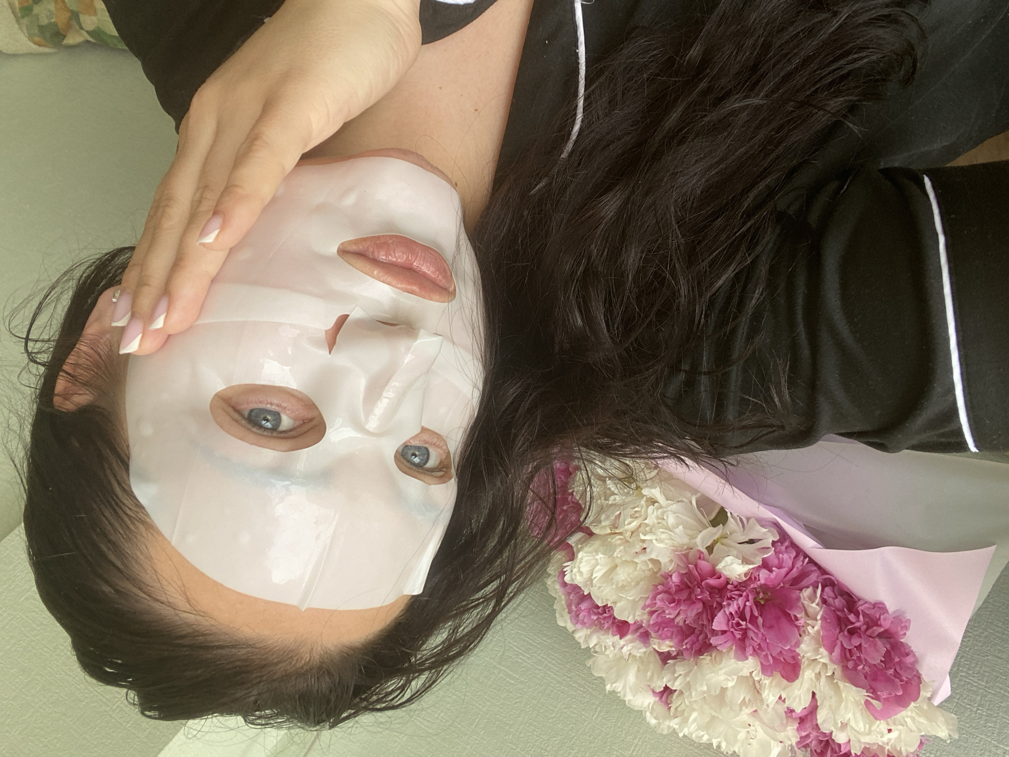 A woman with a white facial mask is lying down, with a bouquet of white and pink flowers beside her. Her dark hair is partially spread around her, and she is wearing a black top with white piping. One hand is raised near her face, showing a French manicure. The background appears to be a light-colored wall. The image does not include enough information to confirm the location in the city of Sochi, Russia.