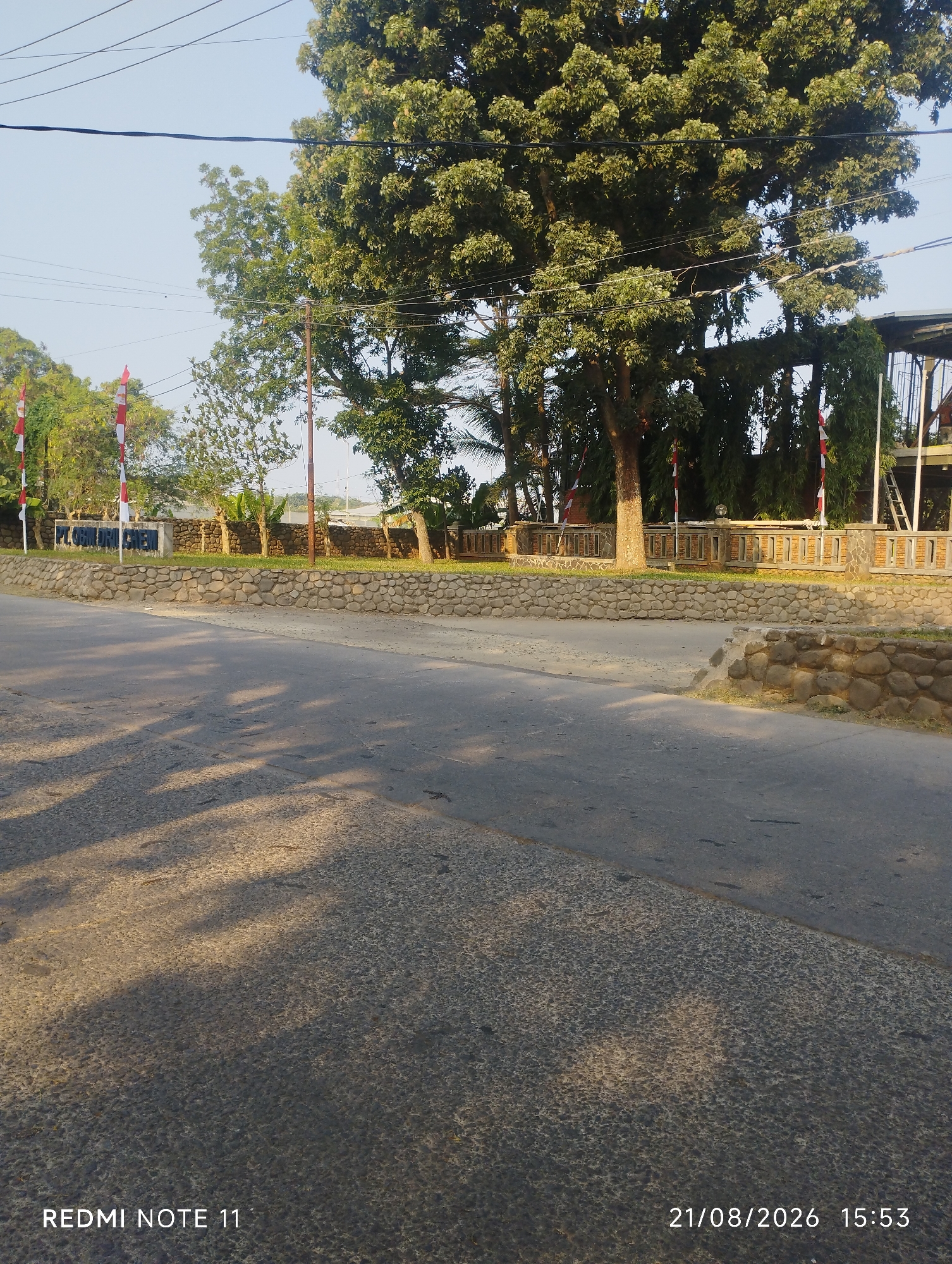 The image captures a roadside scene with lush greenery and a clear sky, likely taken in Karawang, Indonesia. The foreground shows a paved road with distinct tire tracks and shadows cast by trees, indicating a sunny day. Beyond the road, a stone wall borders a grassy area. In the middle ground, a large tree dominates the scene, its branches reaching towards the sky. Several Indonesian flags are visible, planted along the roadside and near a fence. The background reveals a building with a sheltered outdoor area, a ladder, and more foliage. The overall impression is one of a tranquil, possibly semi-urban or rural environment. The bottom of the image displays "REDMI NOTE 11" and "21/08/2026 15:53", suggesting the device and time of capture.