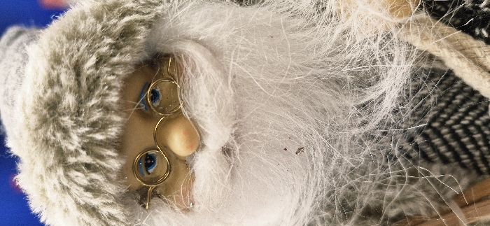 The image provides a vertical, close-up view of a static decorative figurine, located in Vendin-le-Vieil, France. The subject depicts the head and upper body portion of a figure characterized by a light beige face with painted blue eyes accented with white glitter, a small, peach-toned nose, and circular, gold-colored spectacles positioned over the eyes. The figure wears a voluminous, white artificial fur beard, extending from below the nose and covering the lower face and upper chest area. Fine, translucent, hair-like fibers are visible across the beard and spectacles. A grey artificial fur hat, featuring a distinct lighter grey band, is worn on the head. Below the beard, a garment with a black and light grey or white chevron pattern is partially visible. A light beige, coarse-textured braided rope runs vertically along the lower right edge of the frame. The background is an out-of-focus blue field at the upper left, transitioning to darker tones. Small, reddish-brown particulate matter is embedded within the white fur beard, alongside scattered silver glitter fragments. No human figures are present, and no actions or interactions are depicted.