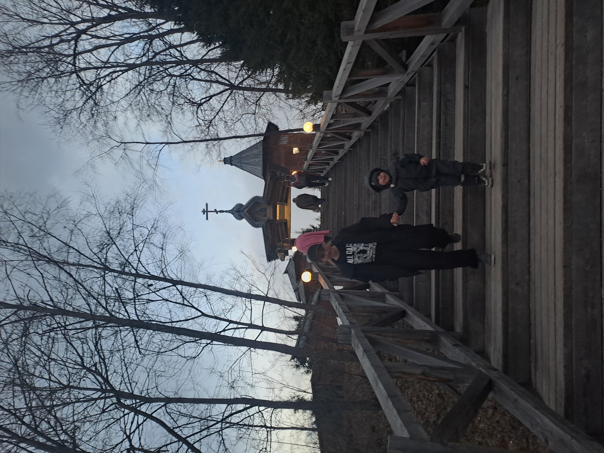 The image depicts a family walking on a wooden bridge leading to a church. The church has a distinct architectural style with a dome and a cross atop it. The sky is overcast, and bare trees flank the bridge. The location is Vzglyadnevo, Russia. The people are on the bridge, walking toward the church.