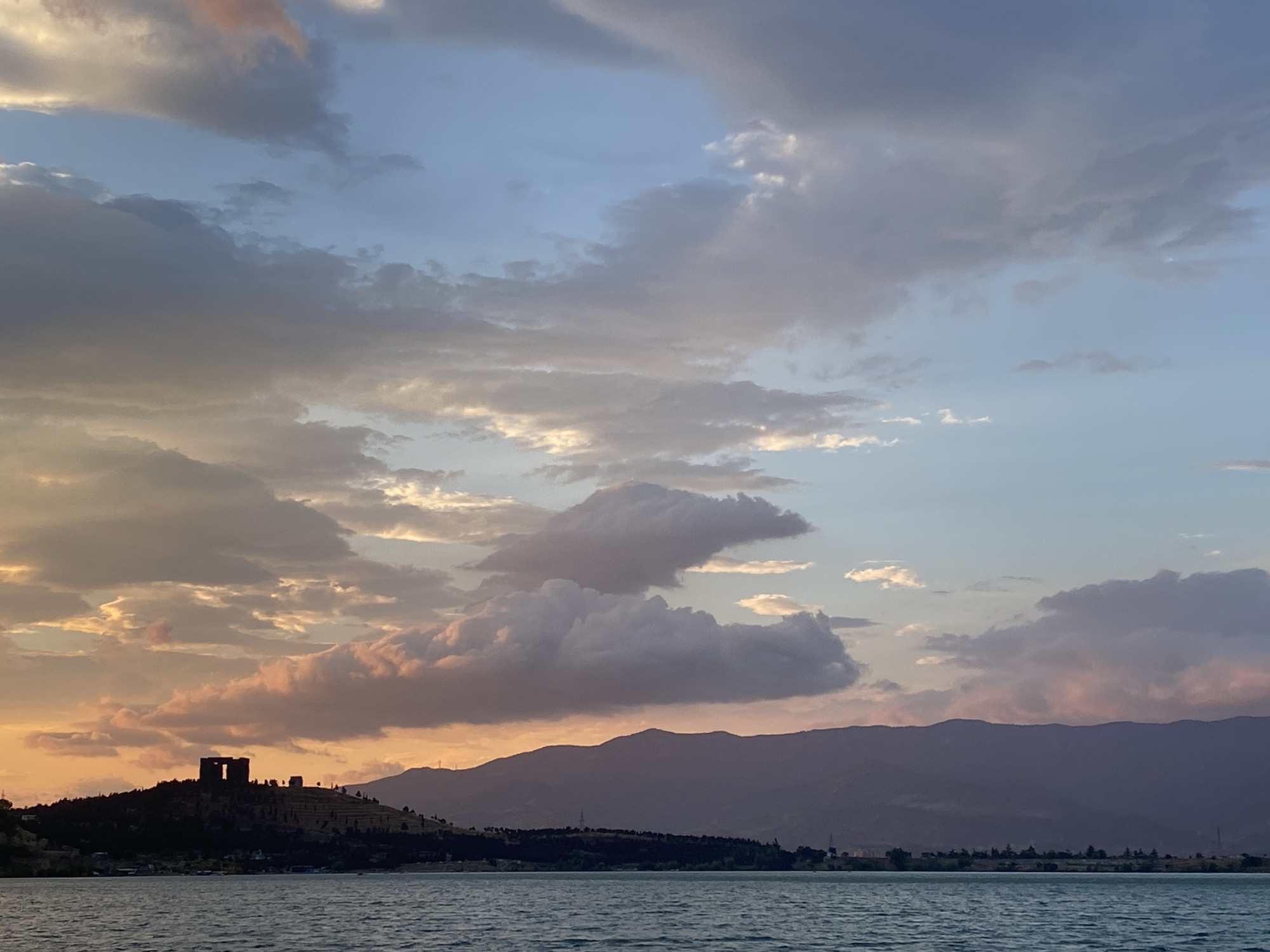 The image depicts a serene waterscape at dusk, with a distant mountainous silhouette dominating the horizon. The sky is adorned with dramatic, cloud-filled formations, transitioning from the warm hues of sunset on the left to cooler blues on the right. In the foreground, a body of water, likely a lake or a wide river, reflects the muted tones of the sky. On the left side of the image, a dark silhouette of a historical structure, possibly a fortress or castle, stands atop a hill, its architectural features partially obscured by the fading light. The hill is dotted with trees and appears to be terraced in some areas. A thin strip of land with more vegetation separates the water from the mountains. The overall atmosphere is tranquil and slightly melancholic, characteristic of twilight. The scene is unlikely to be in Tbilisi, Georgia, as there is no distinct landmark or geographical feature that can be definitively identified as belonging to the city.