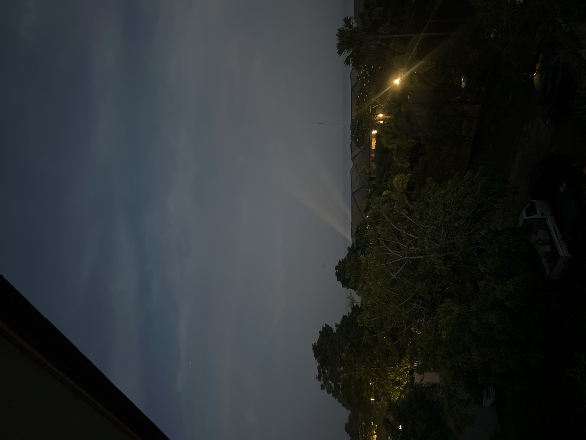 Here is a description of the image:
The image shows a nighttime scene in an urban environment. The sky is overcast with a visible celestial object. There is a line of residential buildings with lit windows. Streetlights illuminate the trees and surrounding area. A car is parked on the side of the road. The location is Unknown, Unknown.