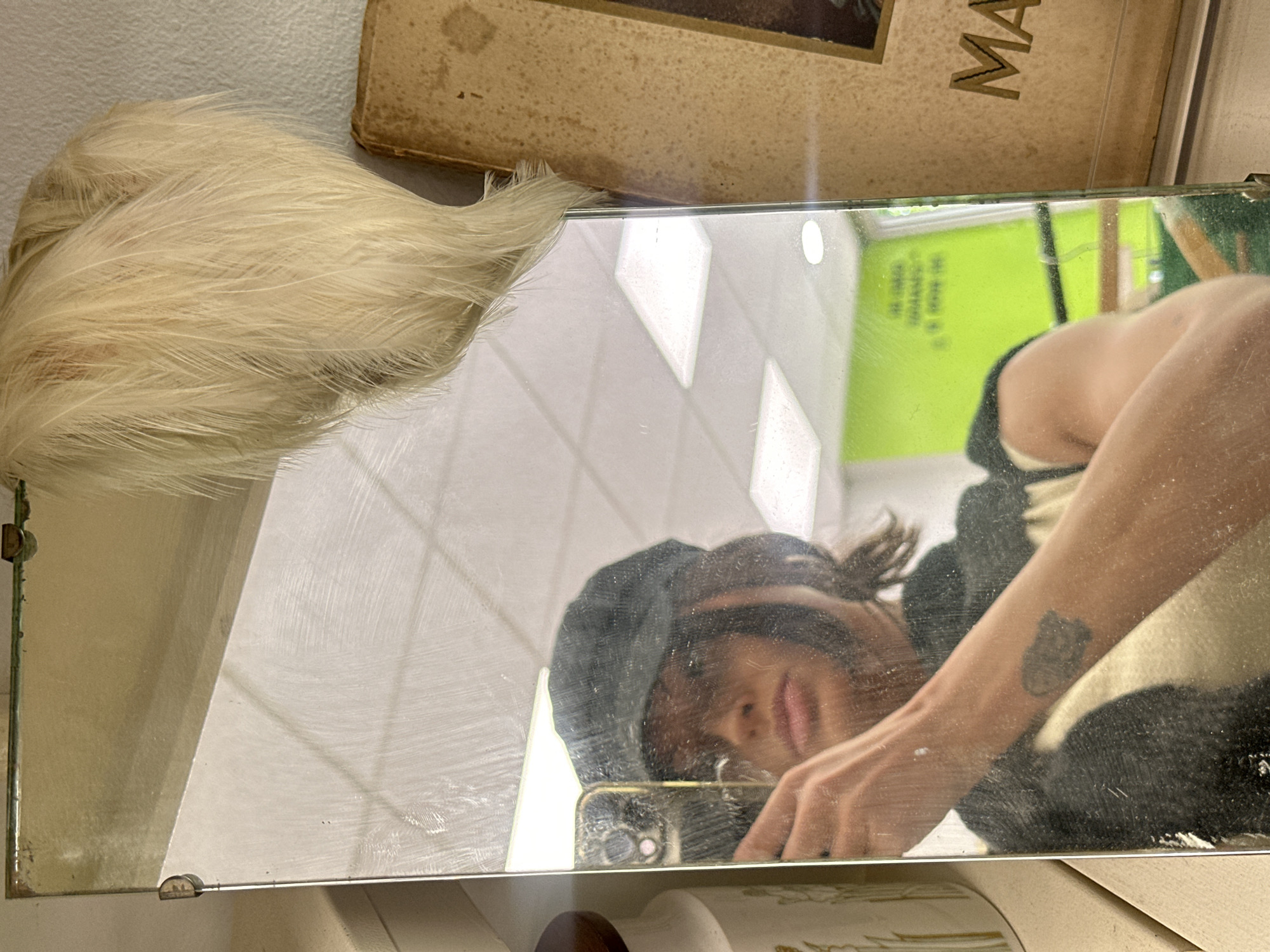 The image is a reflection in a rectangular mirror with tarnished edges. The reflection shows a person, presumably the photographer, holding a smartphone. They are wearing a dark-colored hat and clothing with a light-colored collar. A tattoo is visible on their forearm. Behind the person in the reflection is a bright green wall. To the left of the mirror is a cluster of soft, white feathery material. Above the mirror is a book cover, partially visible, with the letters "MA" in gold. The mirror is propped on a patterned ceramic base. The location appears to be an antique shop in the city of Tunbridge Wells, United Kingdom.