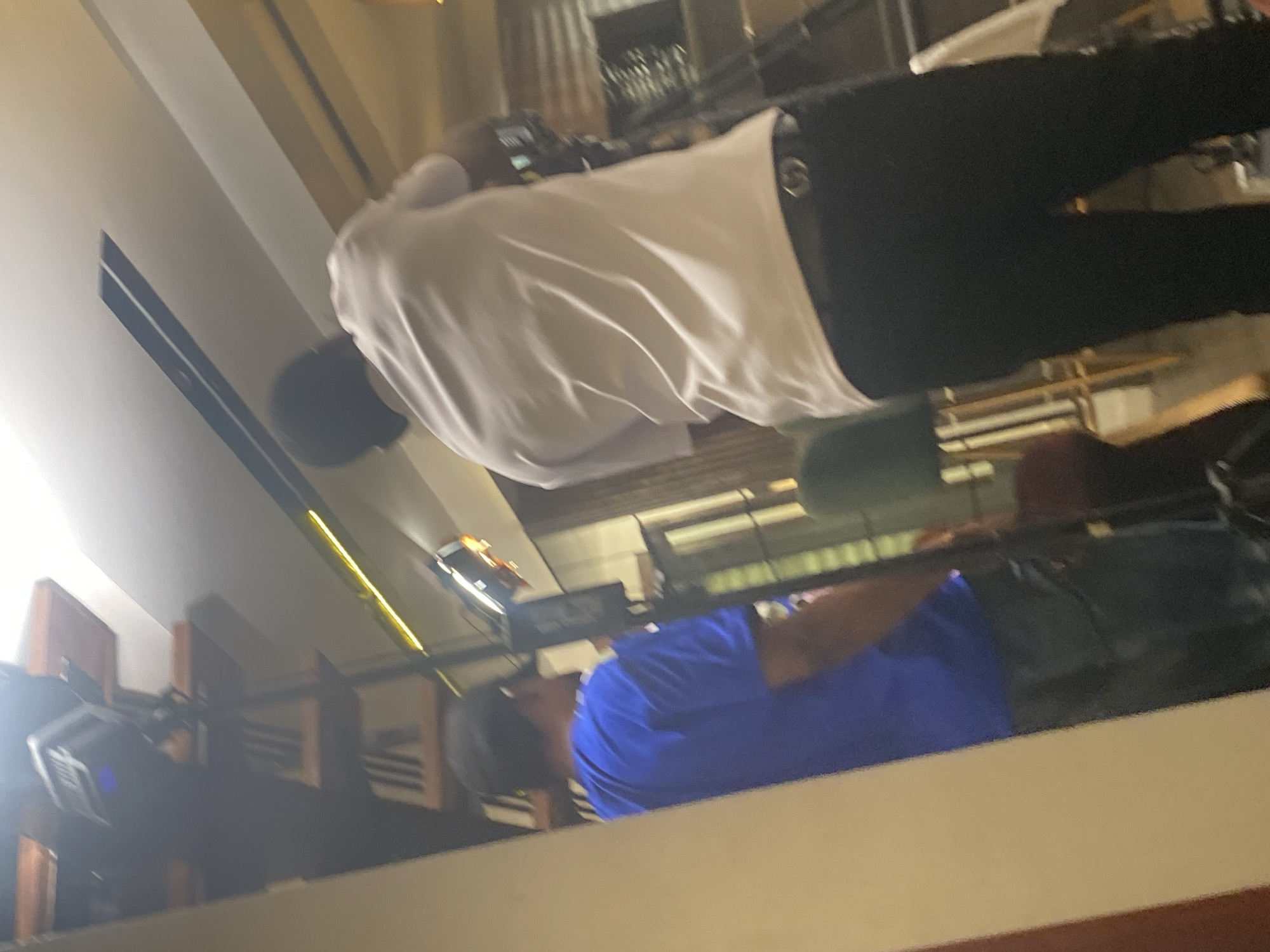 An indoor scene, likely a bar or restaurant, captured from a slightly elevated perspective. The image is rotated 90 degrees clockwise.
In the mid-ground, two individuals are visible, mostly from their backs. The person closer to the foreground is wearing a white long-sleeved shirt, dark pants, and a dark cap. A dark belt is visible at their waist. They appear to be standing upright, facing away from the camera, possibly at a counter or bar. Further back and to the side, a second person is seen wearing a blue short-sleeved shirt and a dark cap. Their upper body is visible, and they are also oriented away from the viewer, seemingly interacting with the same area as the first person.
The foreground features a broad, light-colored surface, possibly a counter or a table edge. The background is characterized by reflections on a dark, polished surface, likely a wall or partition. These reflections show indistinct elements such as wooden panels, linear yellowish light fixtures, and other architectural details. Additional structures, possibly shelving units or bar equipment, are faintly discernible in the deeper background. The lighting is artificial, suggesting an evening or nighttime setting. The overall atmosphere appears casual, possibly in a modern establishment. No visible text is present in the image.