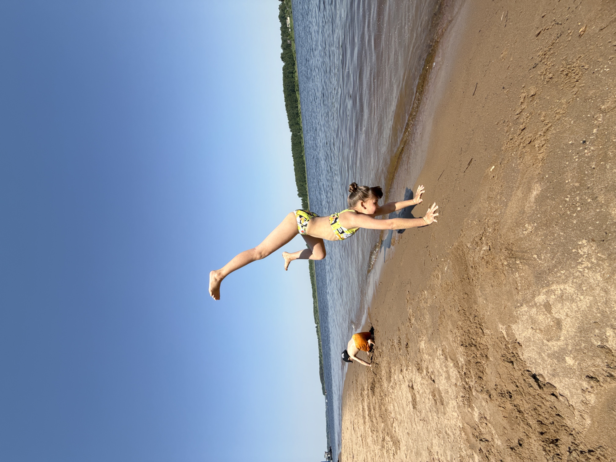 The image captures a scene on a sandy beach adjacent to a body of water, likely a river or lake, and possibly located in the city of Заволжье, Russia. A young woman in a patterned yellow and black swimsuit is depicted in mid-air, in what appears to be a leaping or acrobatic pose, with her hands extended towards the sandy ground. Beside her is another person in orange, digging in the sand. The sky is clear blue, and trees are visible in the background across the water.