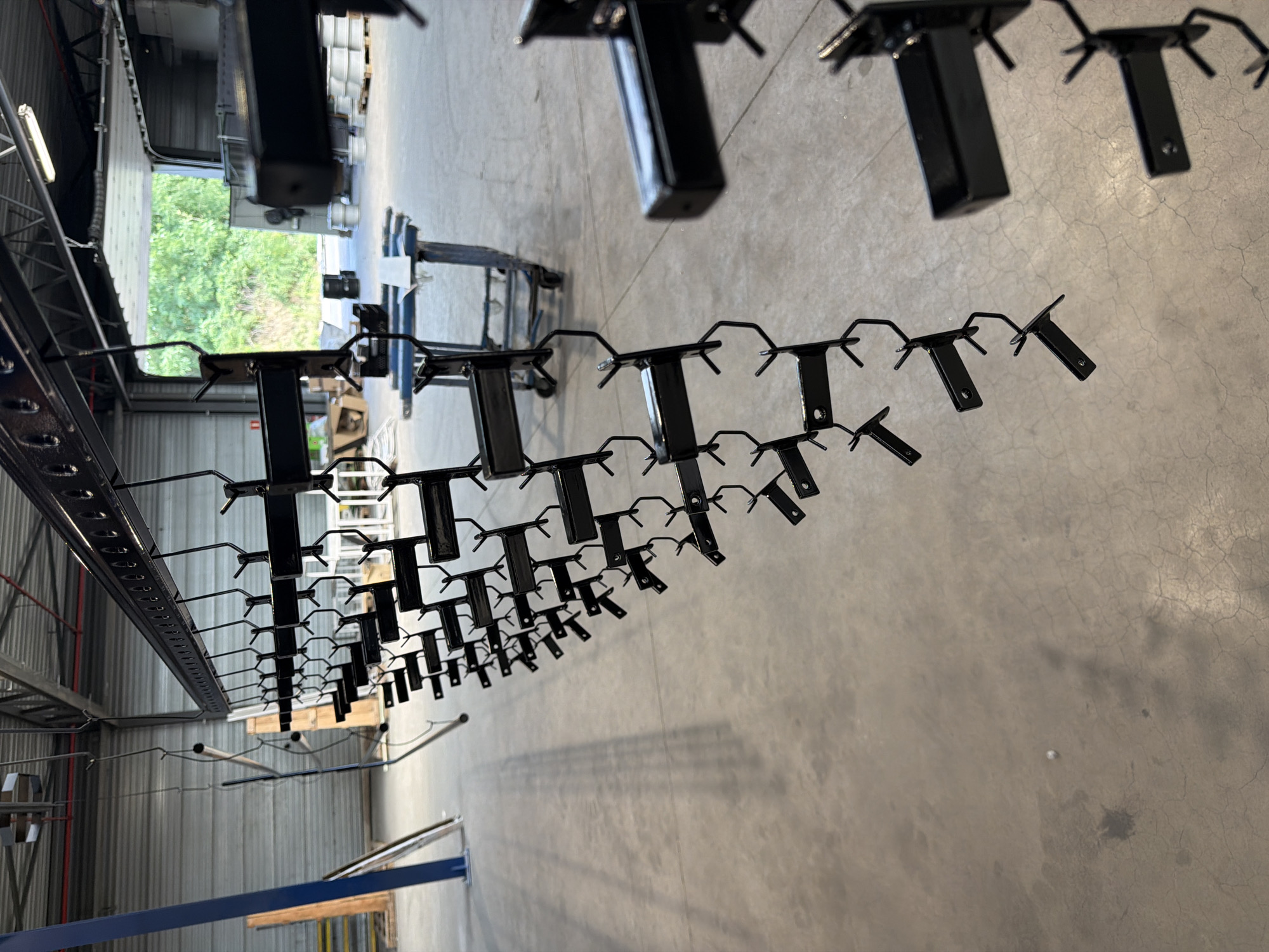 The image shows a close-up, low-angle view of black metal racks and hangers suspended from an overhead track system. The scene appears to be an industrial setting, possibly a warehouse or manufacturing facility, with a concrete floor visible below. The racks are arranged in rows and are designed to hold objects, indicated by the numerous small, hook-like protrusions extending from them. Some of the racks are empty, while others have faint shadows cast on the floor, suggesting they might be in use or have recently been used.
In the background, a large opening reveals lush green trees and foliage, implying the presence of an outdoor area or a large window. The lighting suggests it is daytime, with the light source coming from the open area. The overall impression is one of a functional, organized space, likely involved in production or storage. There are no people visible in the image. The context provided places this scene in Mol, Belgium.