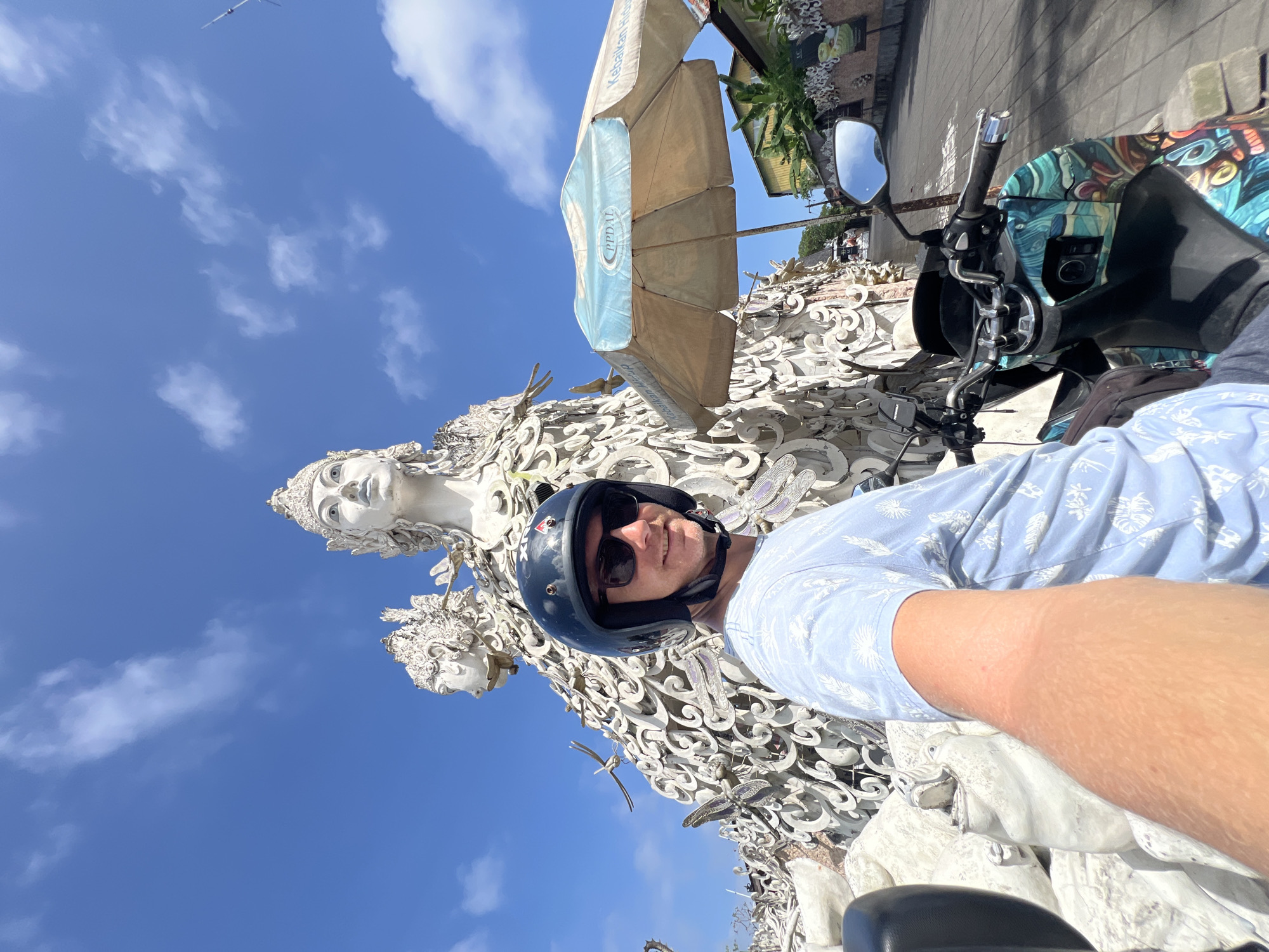 A man wearing a black helmet and sunglasses is taking a selfie while sitting on a blue patterned scooter in front of a large, elaborate white sculpture depicting faces and dragonfly-like figures. The sculpture is situated outdoors under a clear blue sky with scattered white clouds. A beige sun umbrella is partially visible in the background. The location is in the city of Unknown, Indonesia.