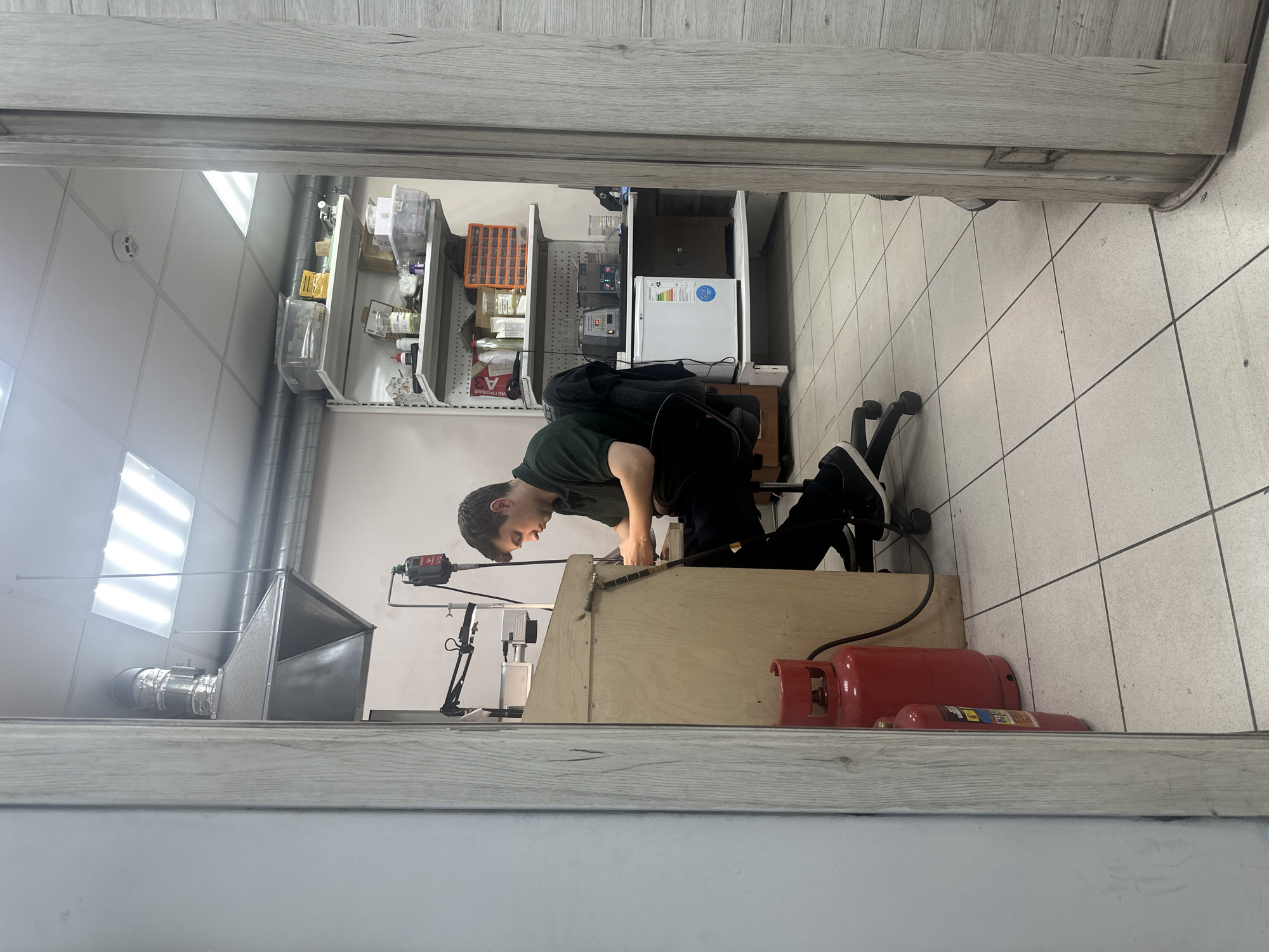 This is a vertically oriented, eye-level photograph taken indoors. The main subject is a young man, dressed in a dark green t-shirt and black pants, kneeling on a black rolling office chair. He is actively engaged in some sort of work or repair, leaning over a wooden workbench. His hands are occupied with tools and equipment mounted on the workbench.
The setting appears to be a workshop or a back room of a store. Behind the man and the workbench, there are shelves stocked with various items, including bottles, containers, and what looks like lab equipment. A small white refrigerator is also visible. To the right of the workbench, there are two red propane tanks. The floor is tiled with light-colored square tiles. Overhead, fluorescent lights illuminate the space. The overall impression is of a functional, somewhat cluttered, work environment.
The image seems to have been taken during daylight hours, judging by the artificial lighting and the lack of external light sources. There are no clear indications of weather or time of day beyond that. The man's expression is focused on his task, suggesting concentration.
Visible text includes what appears to be a label on the smaller propane tank, although it is not legible. A blue sticker with a circular symbol is on the refrigerator.
The location context provided is Tver, Russia.