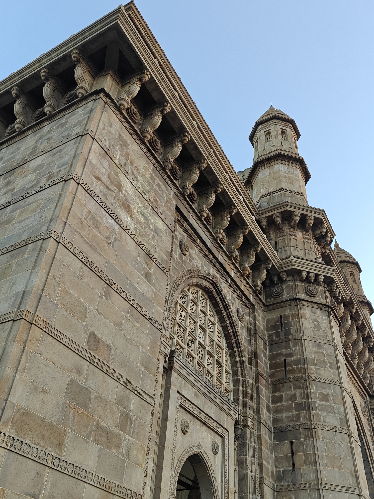 The image shows a detailed view of the Gateway of India, a monument located in Mumbai, India. The perspective is from below, looking up at the structure. The monument is constructed of yellow basalt and reinforced concrete. The stonework includes intricate carvings, patterns, and details. The sky is visible in the background and is a clear, pale blue. There are no people visible in the scene.