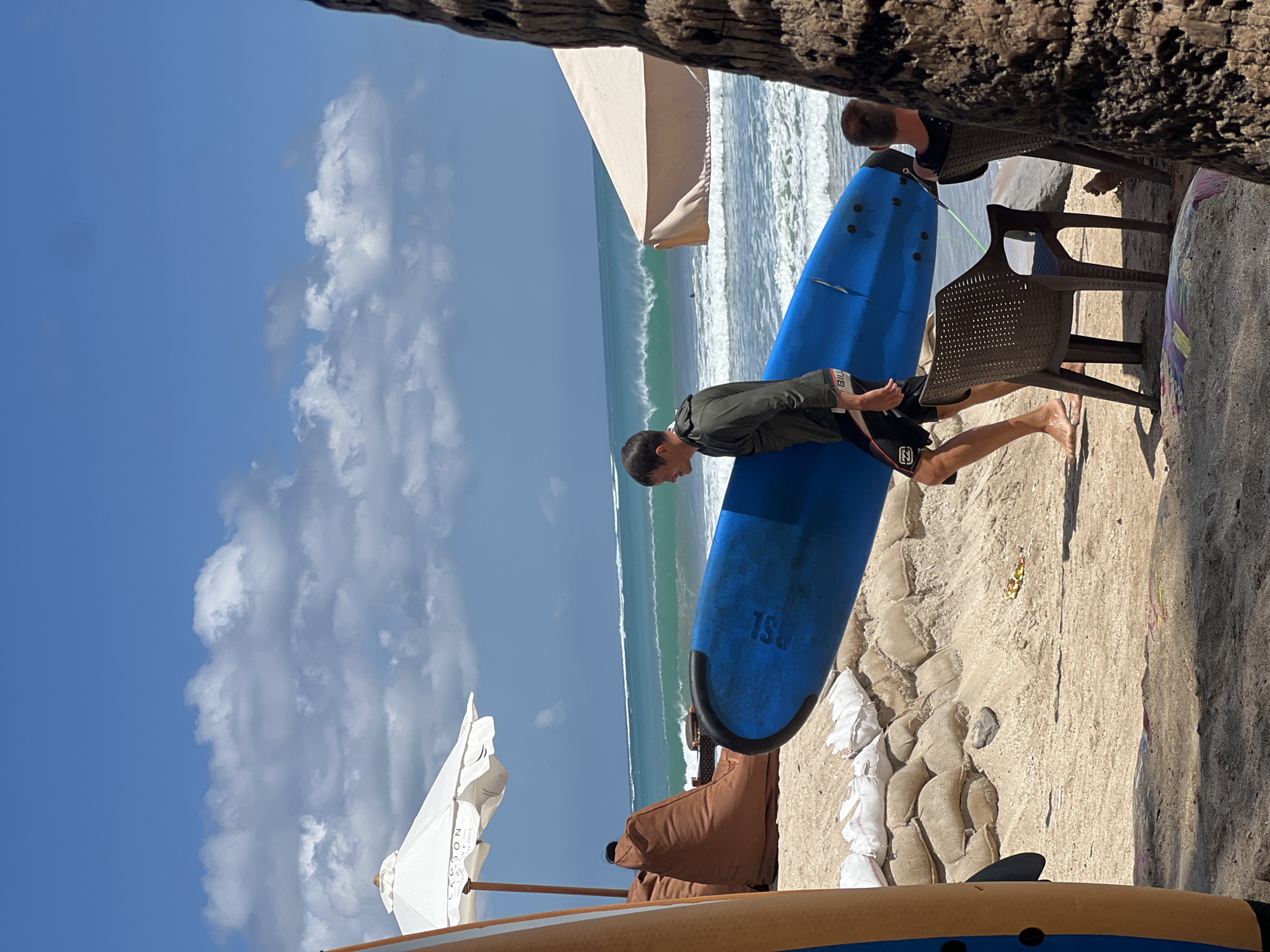 An outdoor, eye-level shot captures a man carrying a blue surfboard on a sandy beach in Kuta, Indonesia. The man, wearing a dark green hooded jacket and black swim shorts, walks from left to right across the frame, his body angled towards the ocean. The surfboard is held diagonally, its tail pointing away from him. In the foreground, a portion of another surfboard, orange and brown, is visible on the left. A brown beanbag chair and a white umbrella are situated on the sand behind the man. The ocean in the background features rolling waves under a bright blue sky dotted with fluffy white clouds. Two plastic chairs, one empty and one occupied by a person facing away from the camera, are in the mid-ground to the right of the man. The scene is characterized by the sandy terrain, the presence of surfboards, and the backdrop of the ocean, suggesting a surfing or beach resort environment.