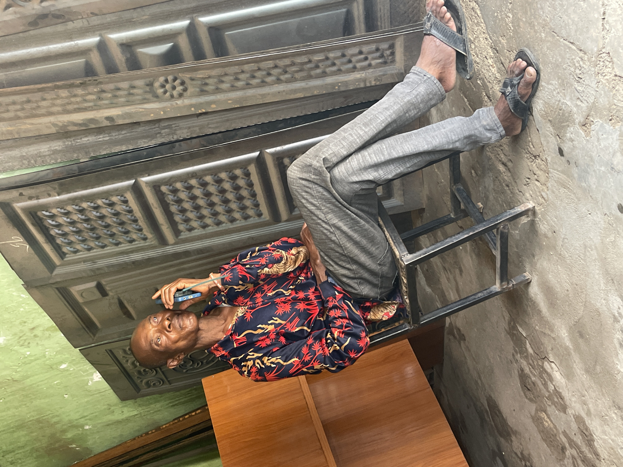 An elderly Black man is sitting on a metal chair inside what appears to be a shop or workshop. The man is light-skinned, balding, and is wearing a red, black, and gold patterned shirt, gray pants, and black sandals. He is holding a blue smartphone to his ear, suggesting he is in the middle of a phone call. Behind the man are multiple gray doors with intricate detailing. To the left of the man is a green wall, and to the right is a gray concrete wall. Underneath the man, is a wooden table. The photo was taken in the city of Zaria, Nigeria.