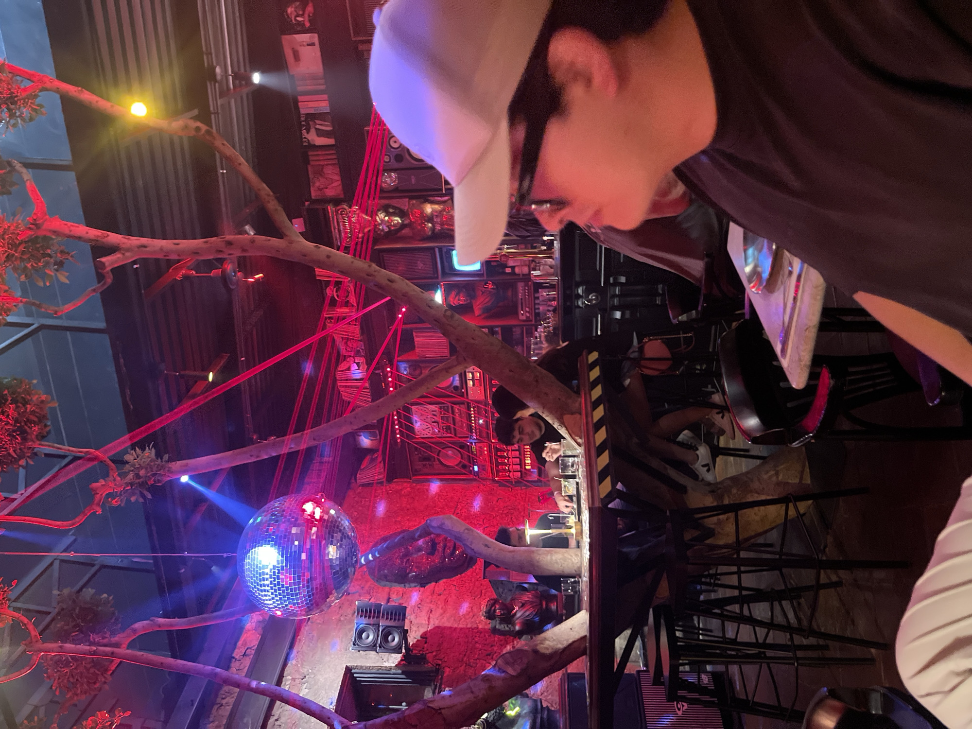 The image captures a dimly lit, indoor setting featuring a disco ball emitting blue light and casting reflections on the walls. A person wearing a white cap and glasses is prominently positioned in the foreground, their face partially visible. Trees and branches are used as a decorative element, adding to the atmosphere. In the background, a stacked display of retro items and electronic devices can be seen. The overall red lighting suggests the ambiance of a nightclub or bar. The location is identified as Puebla, Mexico.