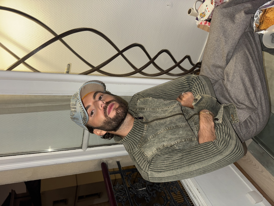 An individual is seated indoors, adjacent to a large window or glass door, with the outdoor night scene of Paris, France, visible to the left. The subject is a male, estimated to be in his 20s or 30s, with dark hair and a beard. He wears a light blue denim baseball cap featuring "MILAN" in studs, a distressed greenish-grey zippered jacket with ribbed sleeves, and grey-brown trousers. His arms are crossed, and a gold-colored bracelet is visible on his right wrist. He faces the camera directly with a neutral expression.
Through the window, a typical Parisian building with light-colored walls and dark windows is discernible under artificial street lighting. A black wrought-iron balcony railing with an ornate design is directly outside the window.
To the right of the subject, an interior wall features a subtle diagonal pattern. A tall, dark decorative lamp with a twisted metal design is positioned against this wall, illuminated from behind. On a surface beneath the lamp, various items are present, including a white mug, several rolls of adhesive tape or fabric, patterned paper or fabric swatches, and a silver foil container holding a pastry-like item. The floor in the foreground is light-colored.