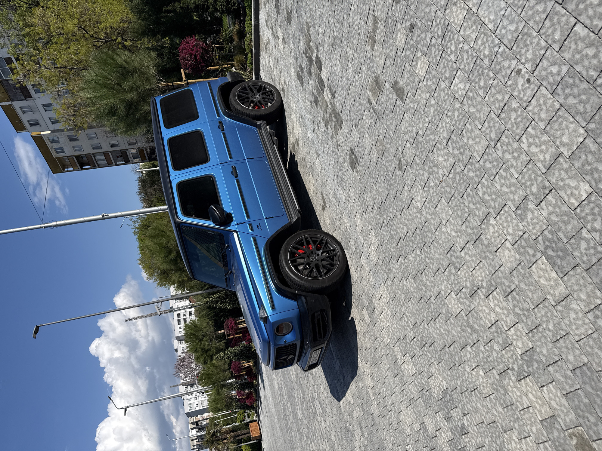A bright blue Mercedes-Benz G-Class SUV is parked on a paved area. The car is angled slightly towards the left of the frame. The paving stones are gray and uniformly arranged. Behind the car, there's a mix of trees, bushes, and buildings. The sky is blue with scattered white clouds. The location is the city of Plovdiv, Bulgaria, as evidenced by the license plate on the car. There are also streetlights visible in the background.