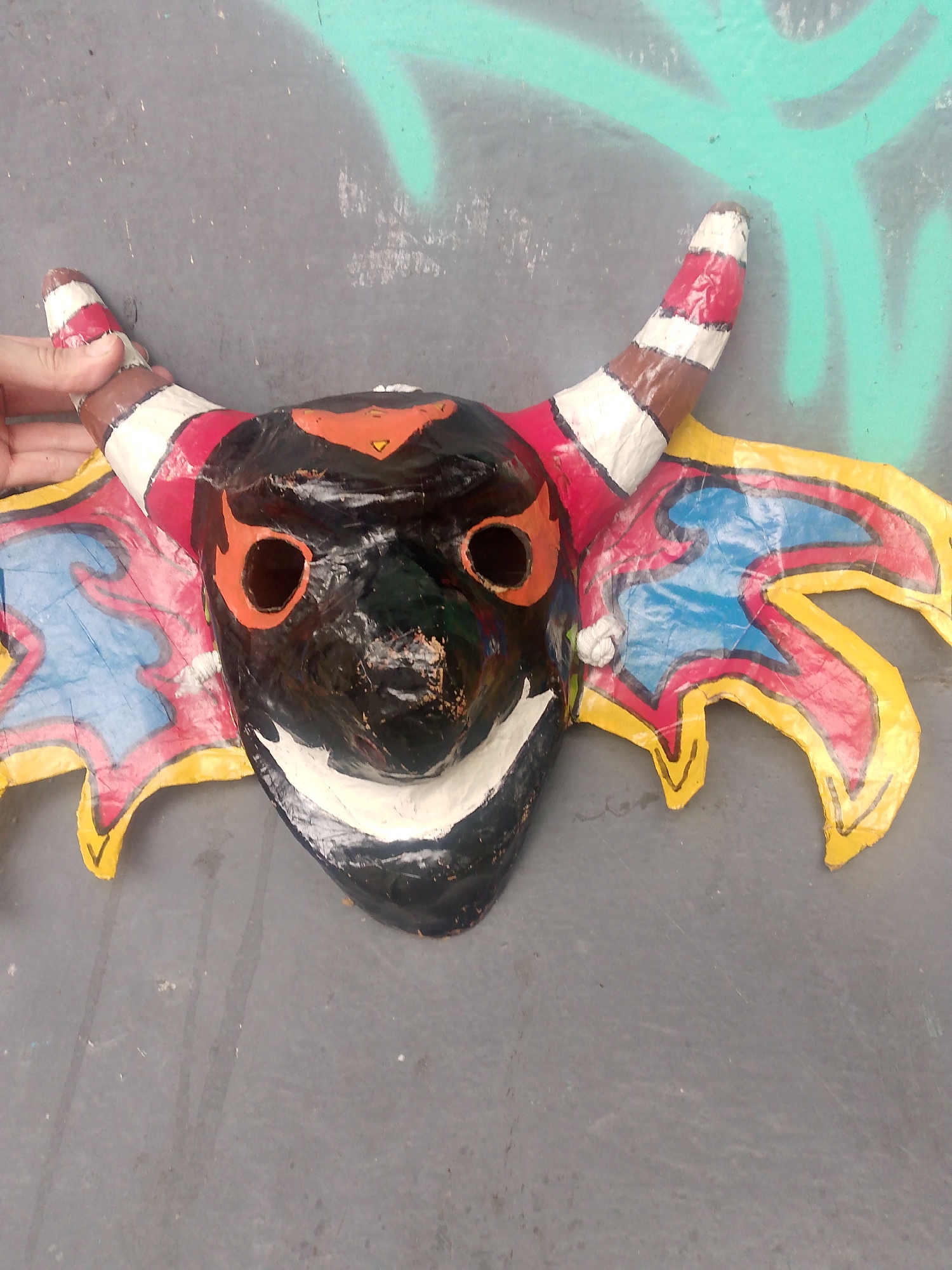 The image depicts a colorful, handcrafted mask lying on a gray surface, possibly concrete. The mask features a black face with orange eye sockets and a small orange triangular shape on the forehead. It has a wide white painted mouth that suggests a smiling expression. Two striped horns, with alternating bands of white, red, and brown, extend upwards. Attached to the sides of the mask are stylized wings, colored with yellow edges, red outlines, and blue and pink designs inside. A hand is partially visible, holding one of the horns. The gray surface has some minor discoloration and possible spray-painted markings in the background. The image was taken in Caracas, Venezuela.