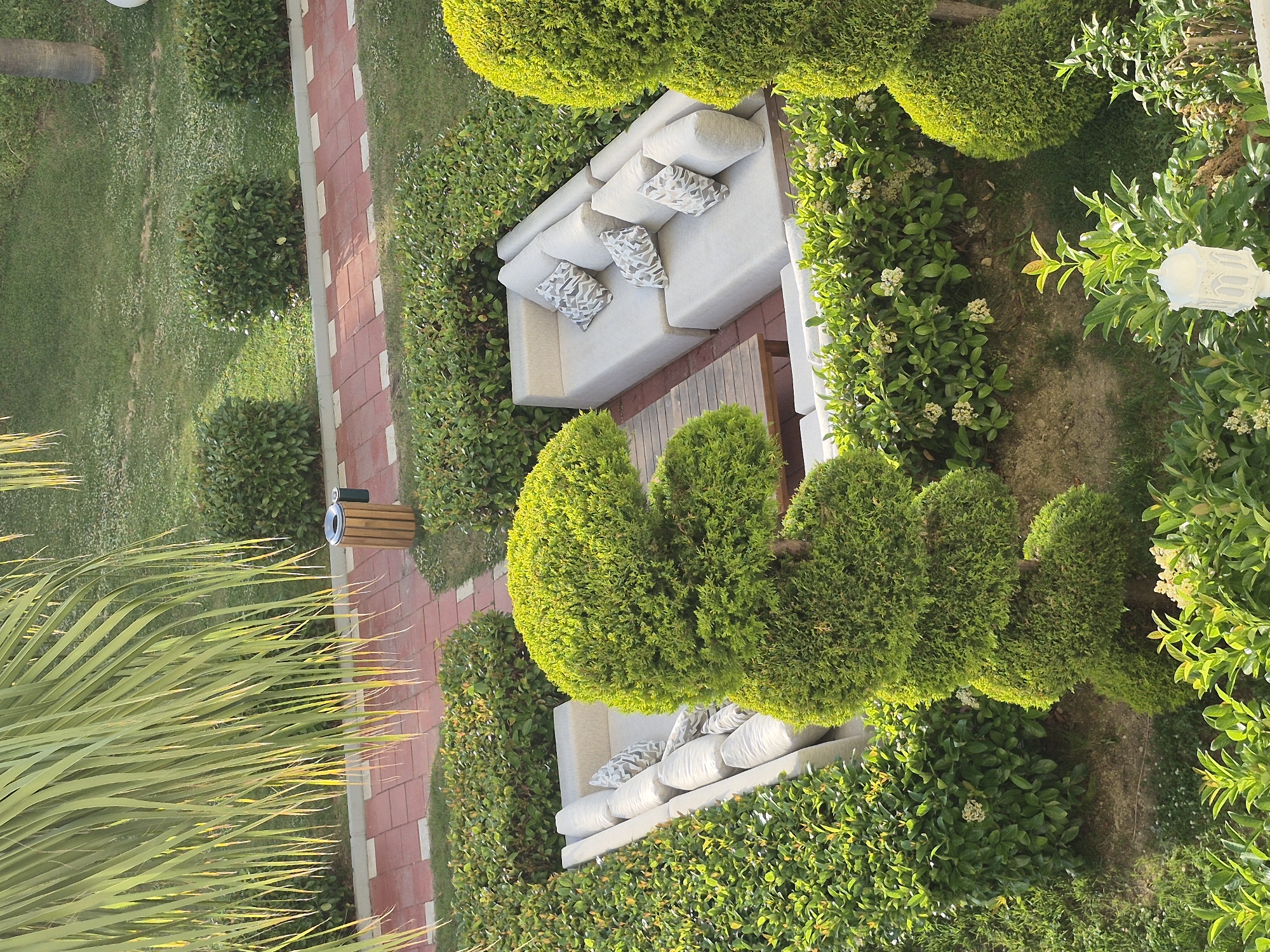 A high-angle shot presents an outdoor lounge area surrounded by lush greenery. Two off-white sofas with patterned throw pillows sit amongst clipped shrubbery. A wooden side table accompanies one sofa. The area is bordered by brick-lined pathways and grassy expanses. Located within Alanya, Turkey. The absence of people or visible human activity suggests a peaceful, unoccupied setting.