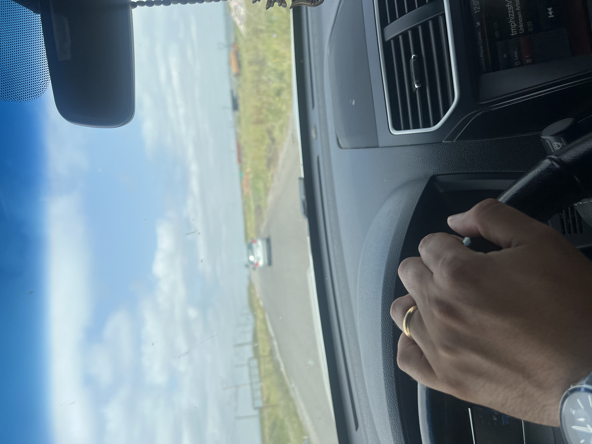 The image shows the interior of a vehicle, looking out through the windshield. A person's hand, wearing a gold ring, is on the steering wheel. The road ahead is paved and appears to be a rural highway. There are cars in the distance, and the landscape on either side is grassy with some industrial structures visible. The sky is partly cloudy. The location is stated to be the city of Jigrasheni, Georgia, though this cannot be visually confirmed from the image itself.