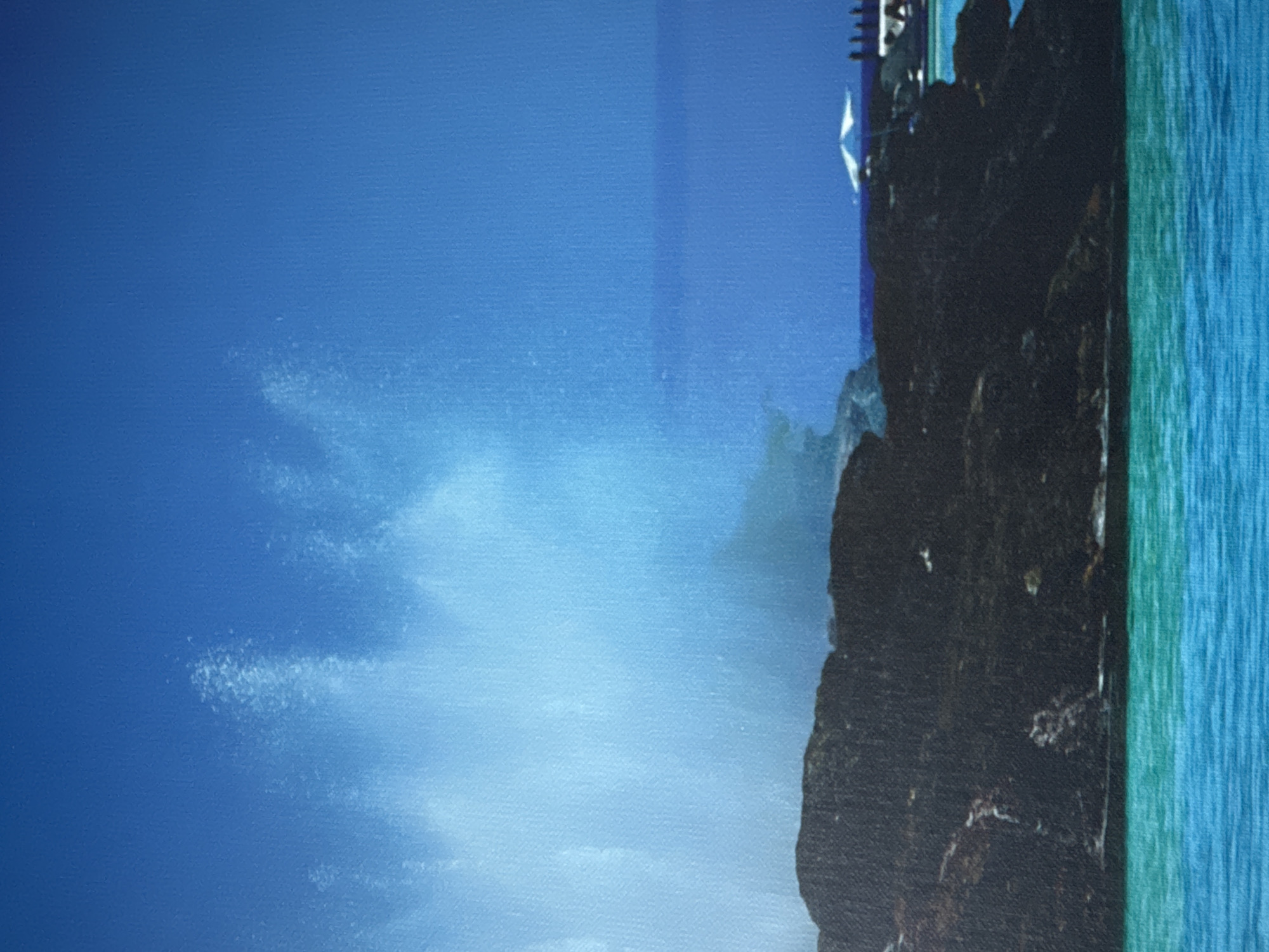 The image shows a dramatic spray of water erupting from a dark, rocky formation along a coastline. The water is a vibrant turquoise, contrasting with the dark rocks and the deep blue of the sky and ocean. In the distance, a white umbrella and a row of silhouetted figures are visible on what appears to be a beach or resort area. The spray of water suggests a natural phenomenon, possibly a blowhole or a wave crashing against the rocks. The scene evokes a sense of powerful natural forces at play. There is no specific indication in the image that this location is in Nesebar, Bulgaria.