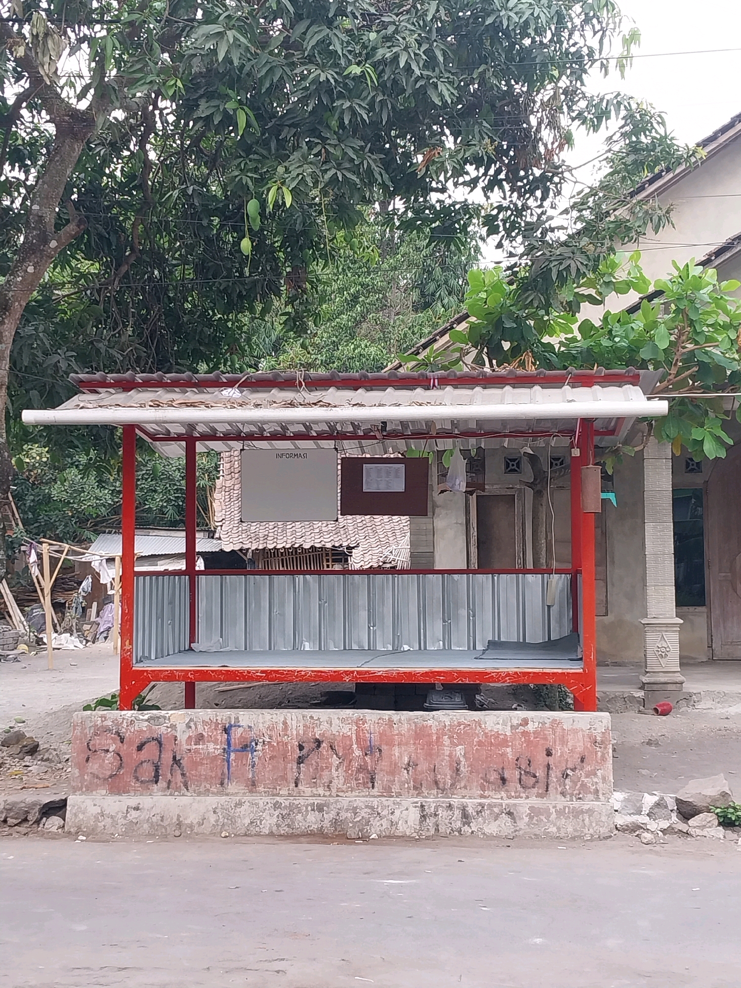 The image shows a small, red-framed shelter with a corrugated metal roof and silver corrugated metal walls. A white whiteboard labeled "INFORMASI" is mounted on the back wall. The shelter sits on a concrete base which is covered in graffiti. In the background, there is a building with a tiled roof and a mango tree with several green mangoes. The setting appears to be a street in Tijayan, Indonesia, indicated by the style of architecture and vegetation. The ground is paved with asphalt and dirt. There are no people visible in the image.