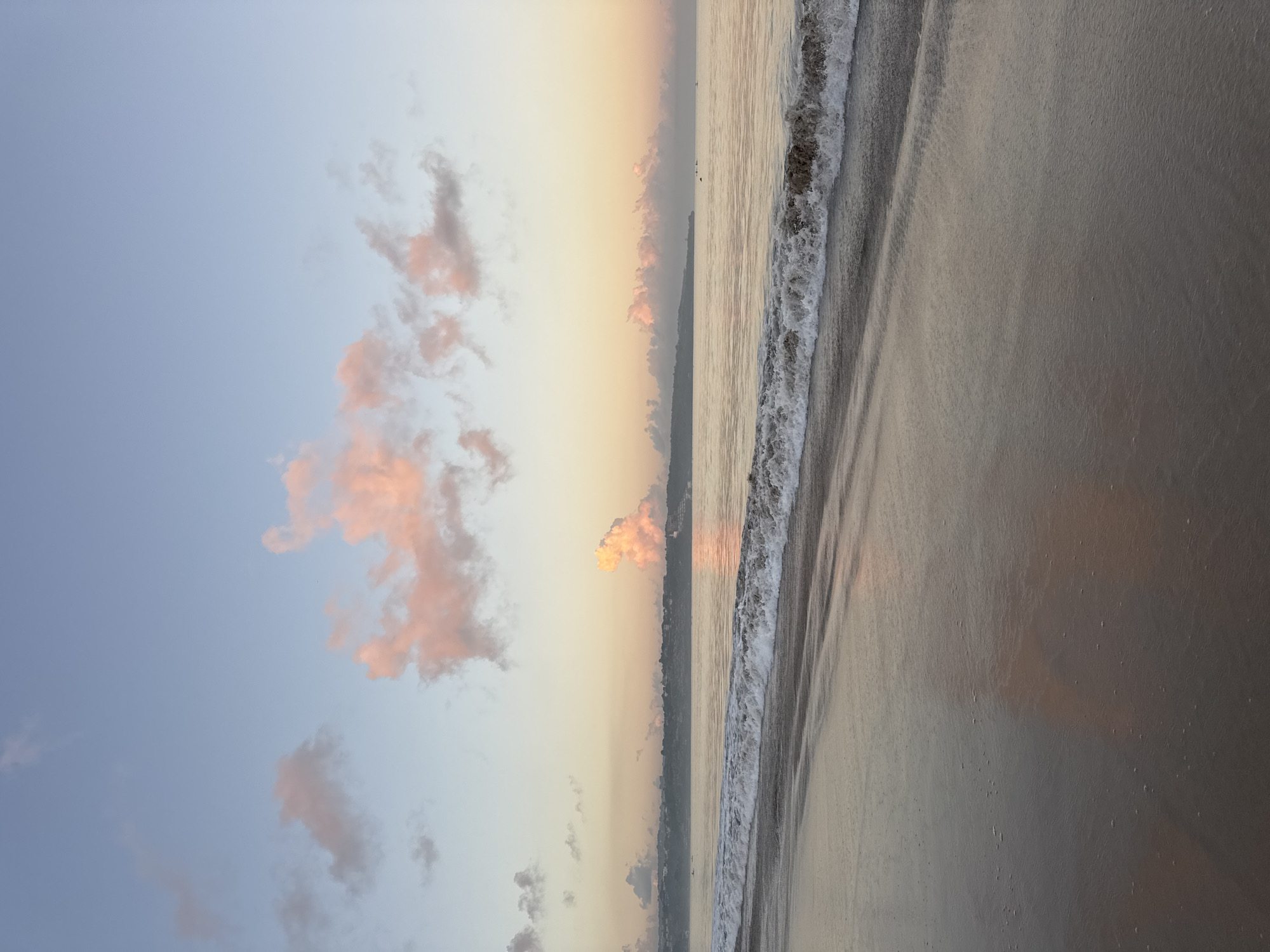 A serene landscape photograph, originally oriented vertically but depicting a horizontal scene, captures a sunrise or sunset at Kuta Beach, Indonesia. The sky dominates the upper portion of the frame, transitioning from a pale blue on the left to soft, warm hues of yellow, orange, and pink towards the right horizon, indicative of early morning or late evening light. Scattered clouds, some tinged with the same rosy and golden tones, drift across the sky.
Below the sky, the calm ocean stretches across the mid-ground, its surface reflecting the soft light. Gentle, foamy waves are seen breaking along the shoreline, creating white crests as they advance onto the wet sand. The expansive sandy beach occupies the foreground, appearing dark and damp from the receding tide, with subtle textures and reflections visible on its surface.
In the far distance, along the horizon where the sky meets the sea, a faint, dark silhouette of land or hills is visible, suggesting a distant coastline. No people or visible text are present in the image, emphasizing the natural beauty and tranquil atmosphere of the beach scene. The overall impression is one of peacefulness and natural grandeur at dawn or dusk.